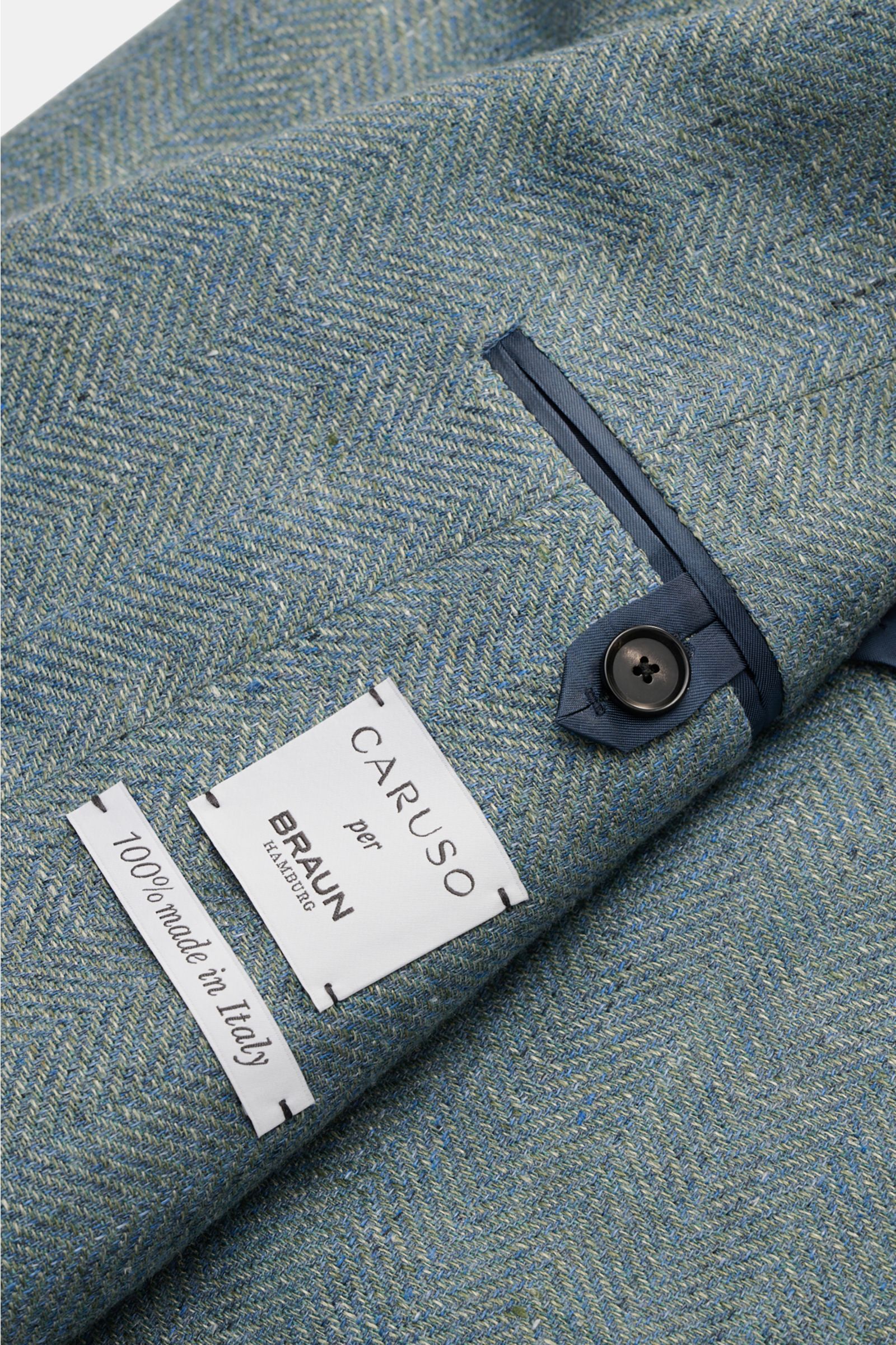 Close-up top view of Caruso Smart-casual jacket 'Aida' grey-green mottled showing the herringbone fabric, buttoned cuff, and brand labels.

Description: In the smart-casual jacket 'Aida', CARUSO combines smart style with fabric in the natural materials