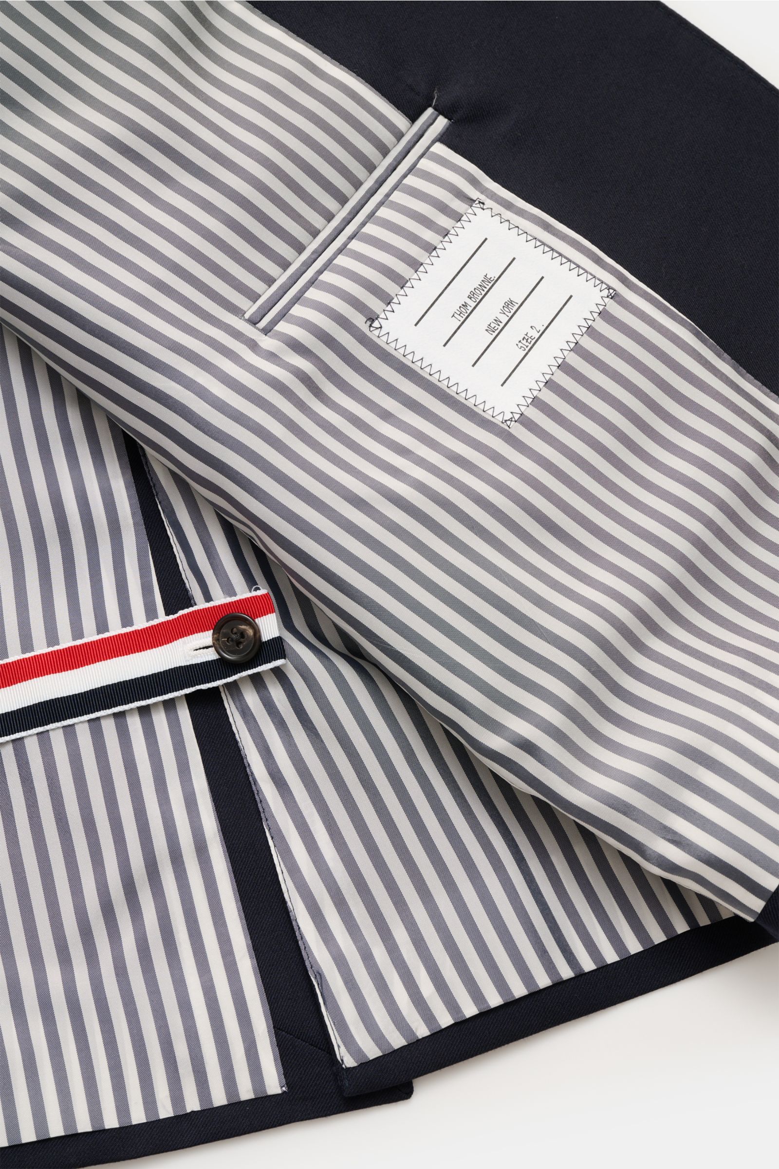 Innenansicht des Thom Browne Sakko navy, fotografiert aus Nahaufnahme schräg von oben, mit gestreiftem Futter, Ripsband in Tricolore und Innentaschen.