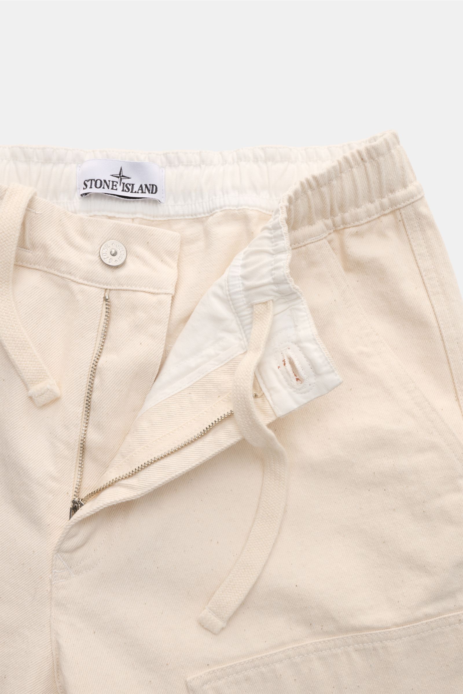 Close-up front view of Stone Island Jogger pants 'Raw Beauty' cream, showing zip, waist button, elasticated waistband, and drawstring.

These robust jogger pants called 'Raw Beauty' by STONE ISLAND are made of distinctive pure cotton twill in workwear sty
