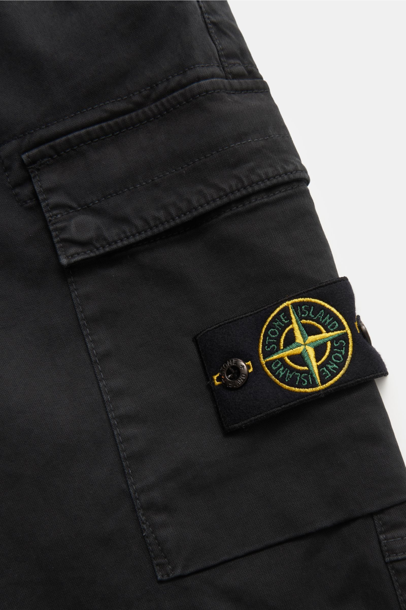 Close-up view of Stone Island Cargo-Joggpants schwarz black cotton fabric showing classic cargo pocket with flap, typical compass badge, and fine stitching details. Features include leicht elastische Baumwolle, weicher Griff, garment dyed, Regular Fit, ku