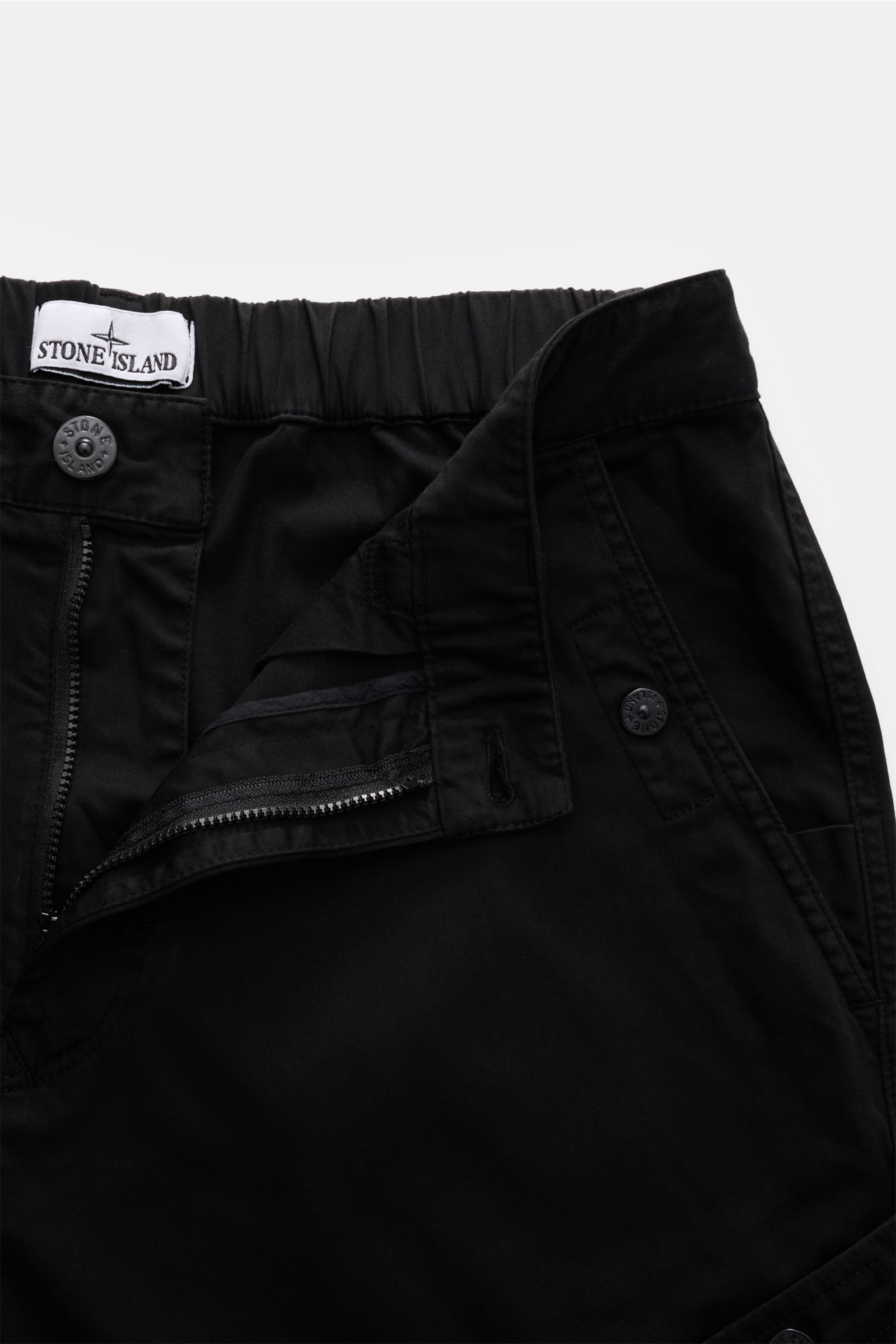 Nahaufnahme der geöffneten Stone Island Cargohose schwarz von oben, mit teilelastischem Bund, Reißverschluss, Druckknopftaschen und Bio-Baumwolle.