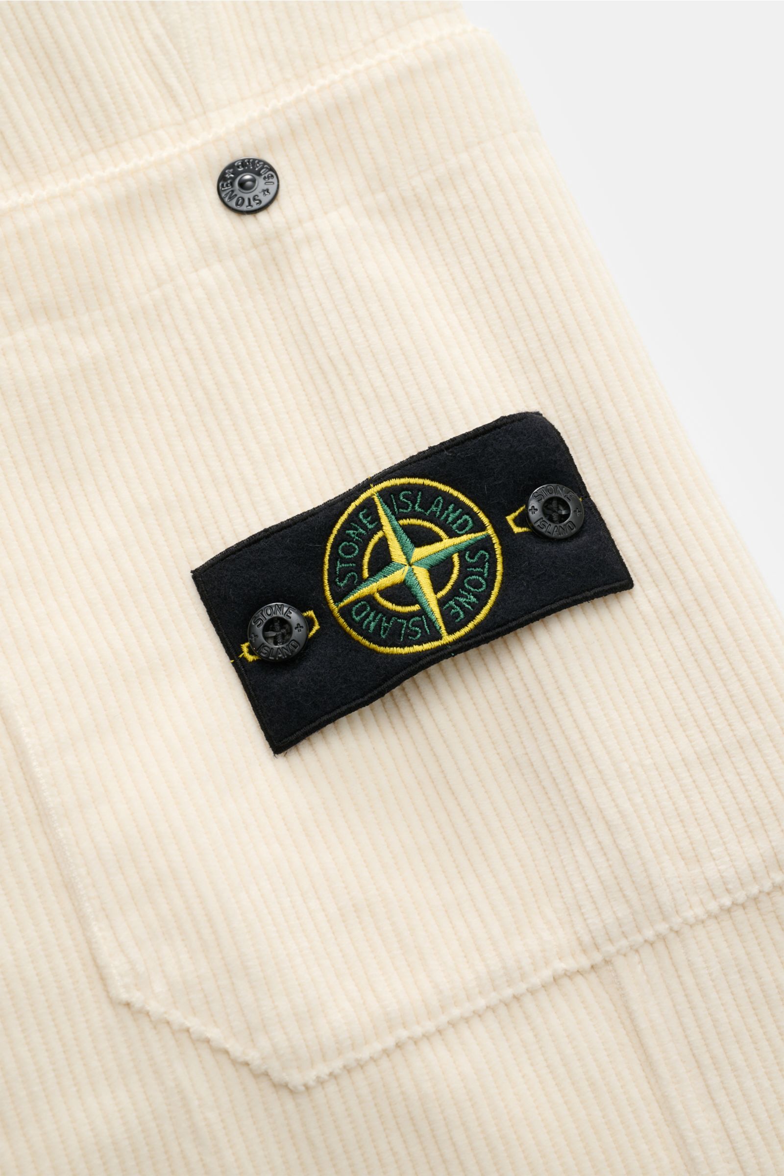 Nahaufnahme der Stone Island Cord-Cargohose creme aus Bio-Baumwolle, Fokus auf Cargo-Tasche mit Druckknopf und Kompass-Badge.