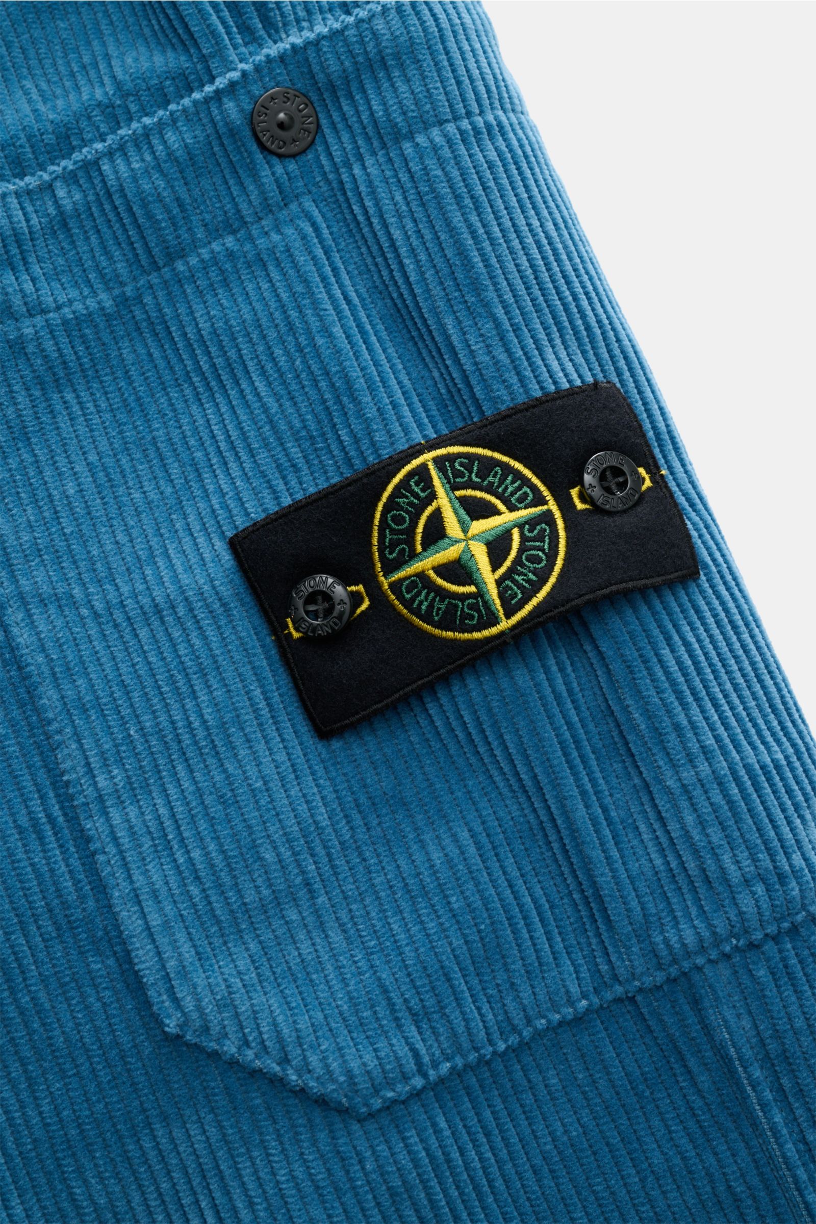 Nahaufnahme der Stone Island Cord-Cargohose blau, Detailansicht der seitlichen Cargo-Tasche aus Bio-Baumwoll-Cord mit weichem Griff und dem ikonischen Kompass-Badge, fotografiert aus schräger Perspektive.