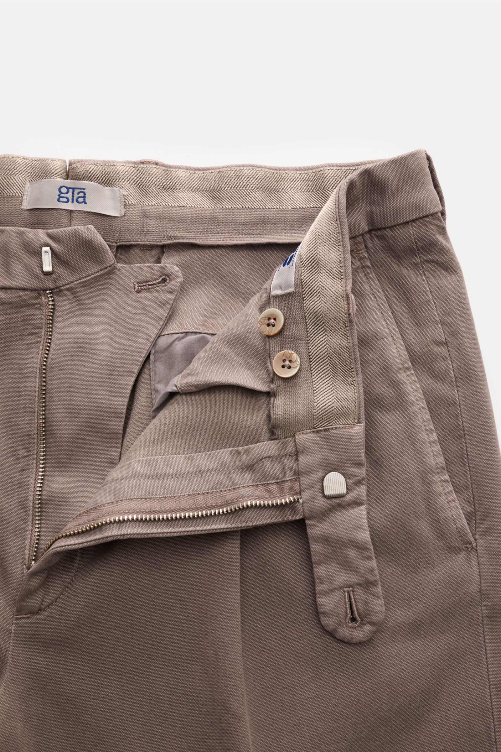 Close-up, top-down view of the GTA Hose 'Luca' taupe showing the zipper, double waistband pleat, elastic waistband insert, button fly, and front pocket with fine details in a soft cotton-modal blend.