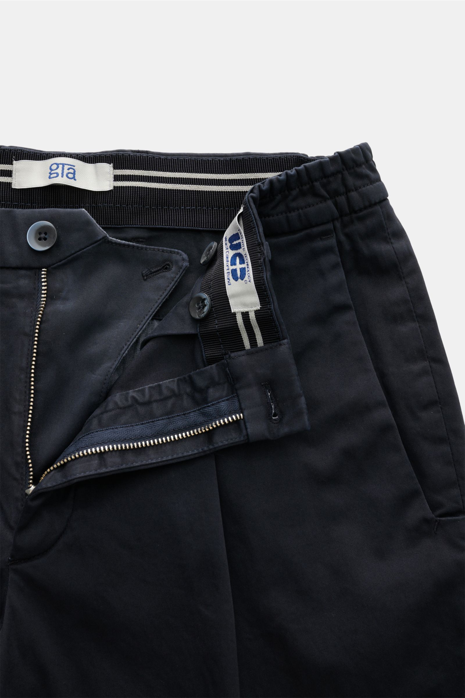 GTA Cotton trousers 'Dennis' navy shown from a close-up top-front view, featuring a partially elasticated waistband, zip, buttons, French pockets, and soft slightly elastic tapered cotton fabric. 

The cotton trousers 'Dennis' by GTA are defined by the
