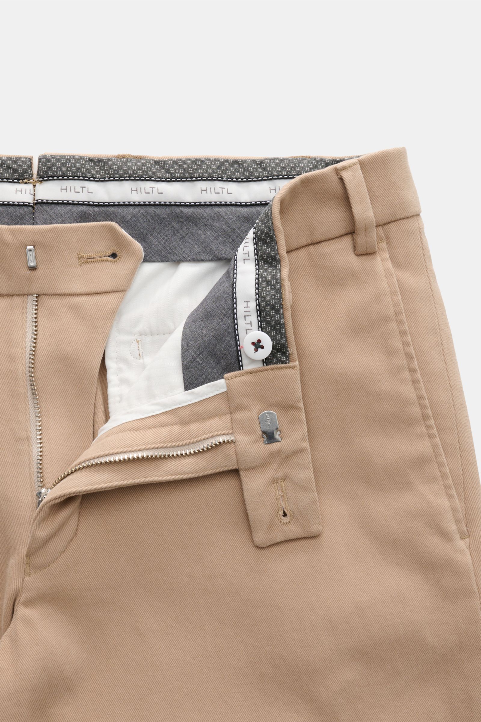 Close-up view of the opened zipper and waistband of Hiltl Baumwollhose 'Parma' sand, showing elastic cotton fabric, soft touch, slim fit, slightly tapered cut, zipper and button closure, French pockets, and welt back pockets with buttons. Vielseitiges Ess