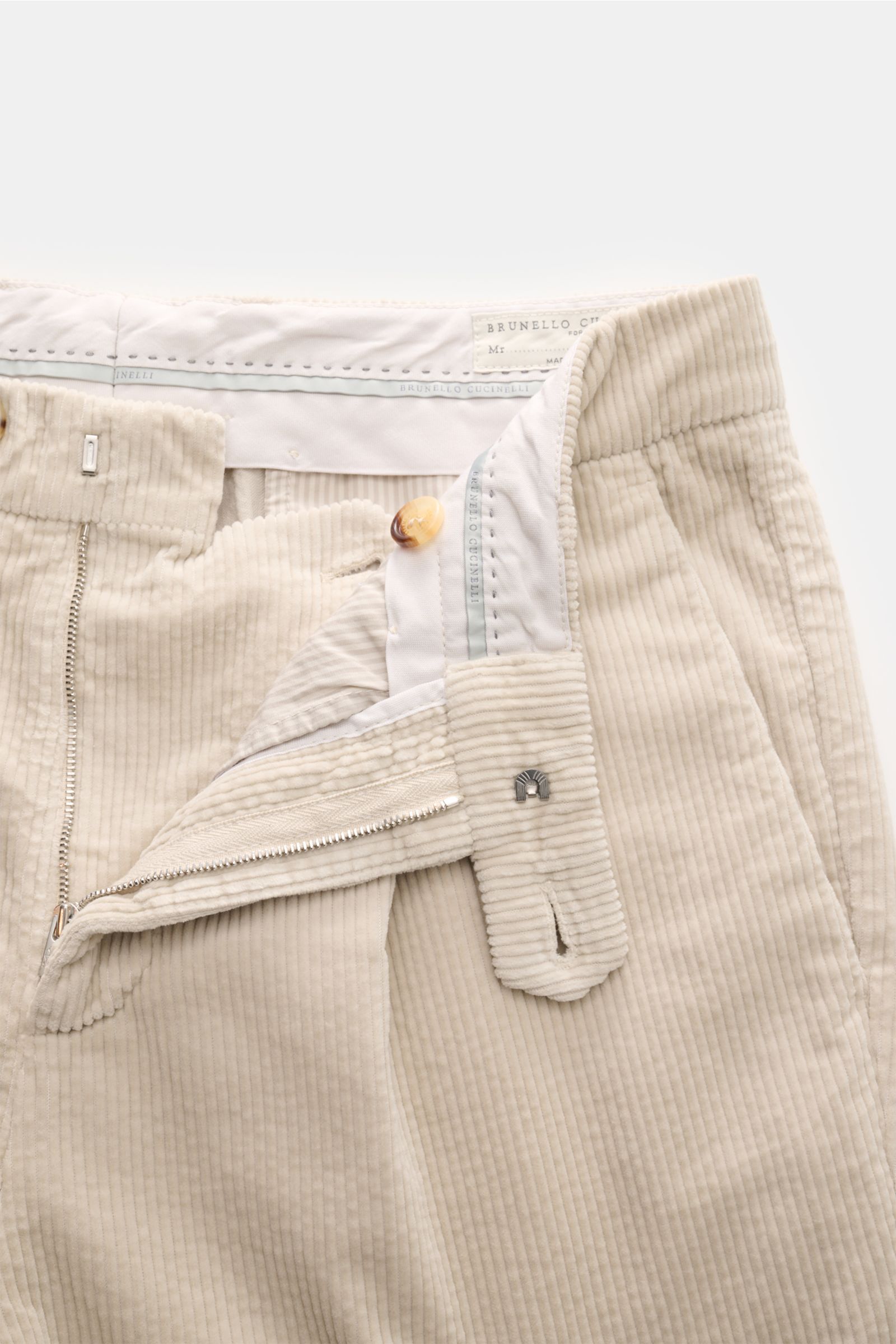 Brunello Cucinelli Cordhose creme shown from a top close-up perspective with zipper open, revealing pure cotton fabric, soft touch, regular fit, very slim taper, zipper and button closure, simple pleat, French pockets, and welted back pockets. Eleganz mit