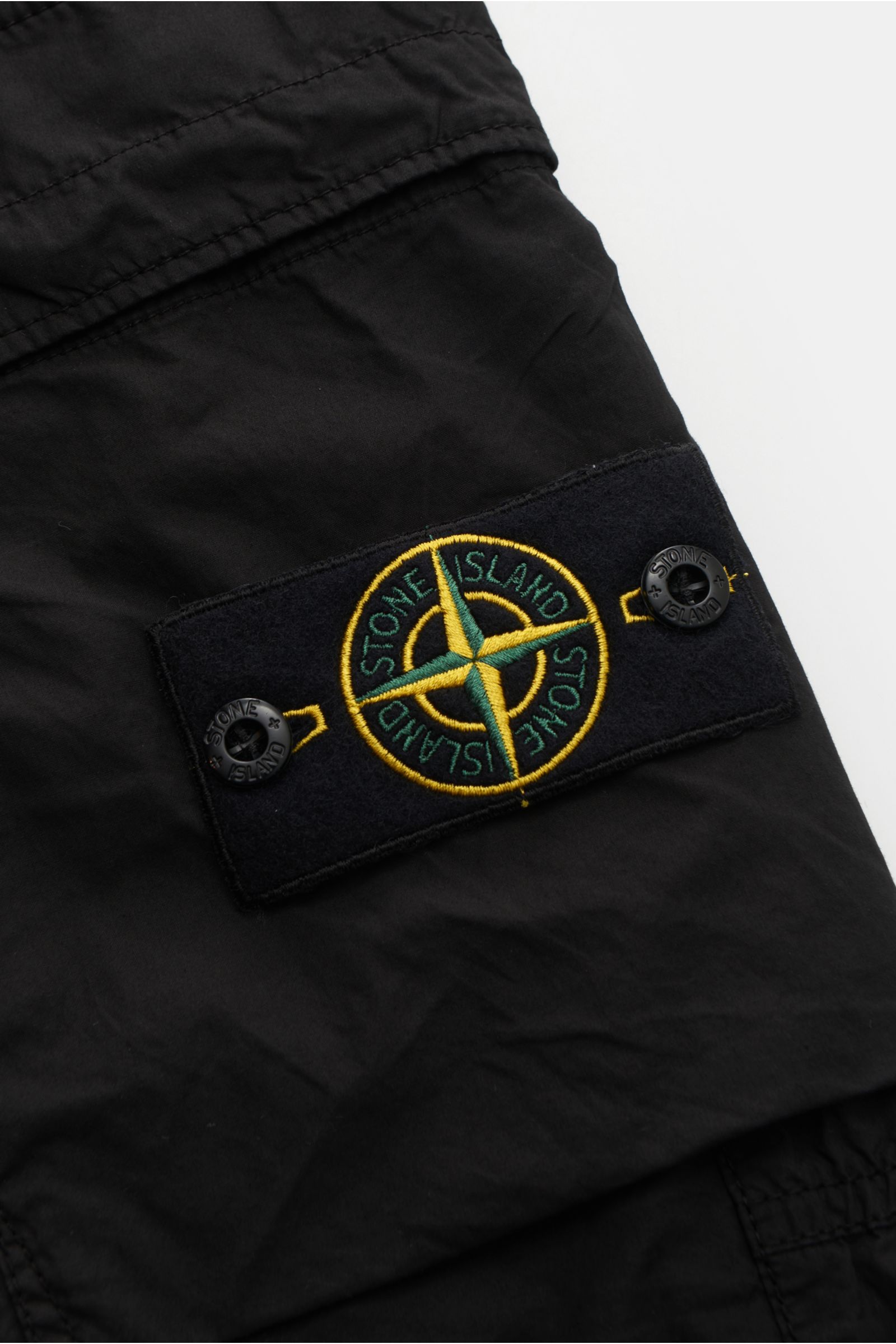 Close-up side view of the Stone Island Cargo-Jogpants 'Regular Tapered' schwarz showing the iconic compass badge on black elastic cotton fabric with a soft touch, featuring classic cargo pockets, elastic waistband with internal drawstring, zippered side p