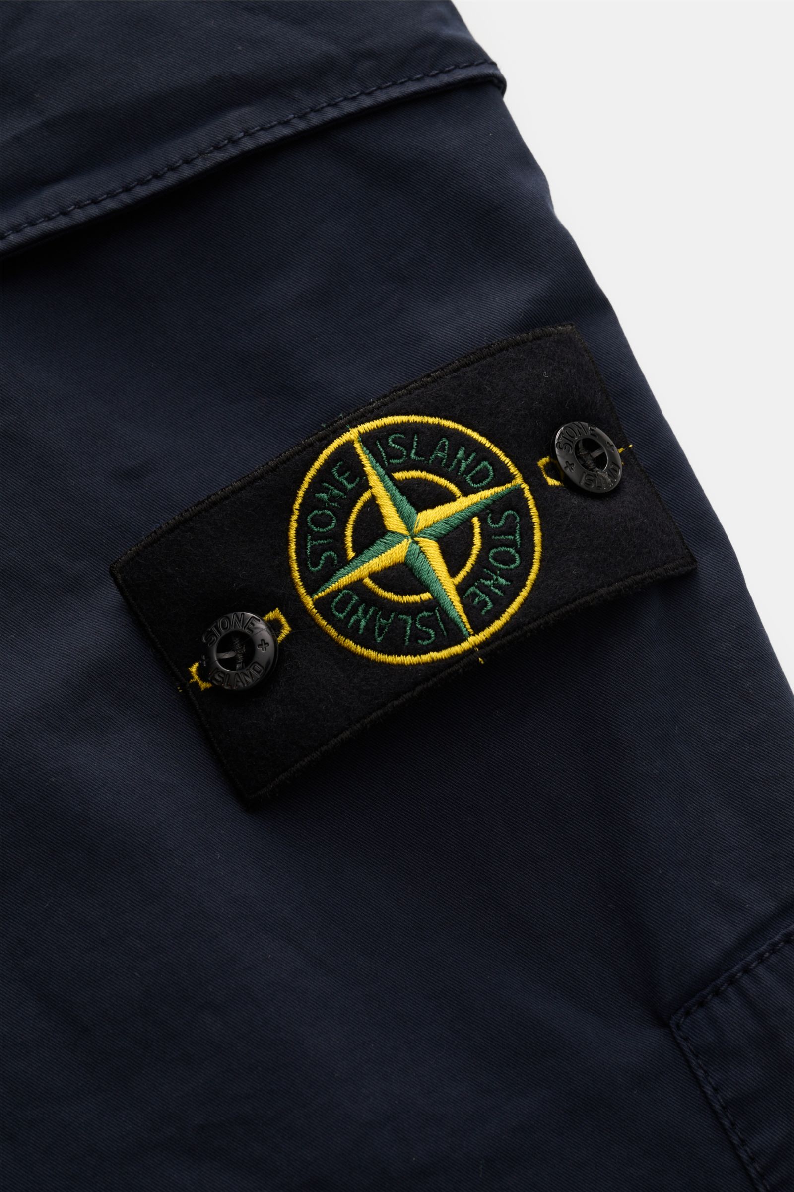 Nahaufnahme der Stone Island Cargohose navy aus Baumwoll-Twill, Detailaufnahme des typischen Kompass-Badges mit Knöpfen, weicher Griff, slim fit, schmal zulaufend.