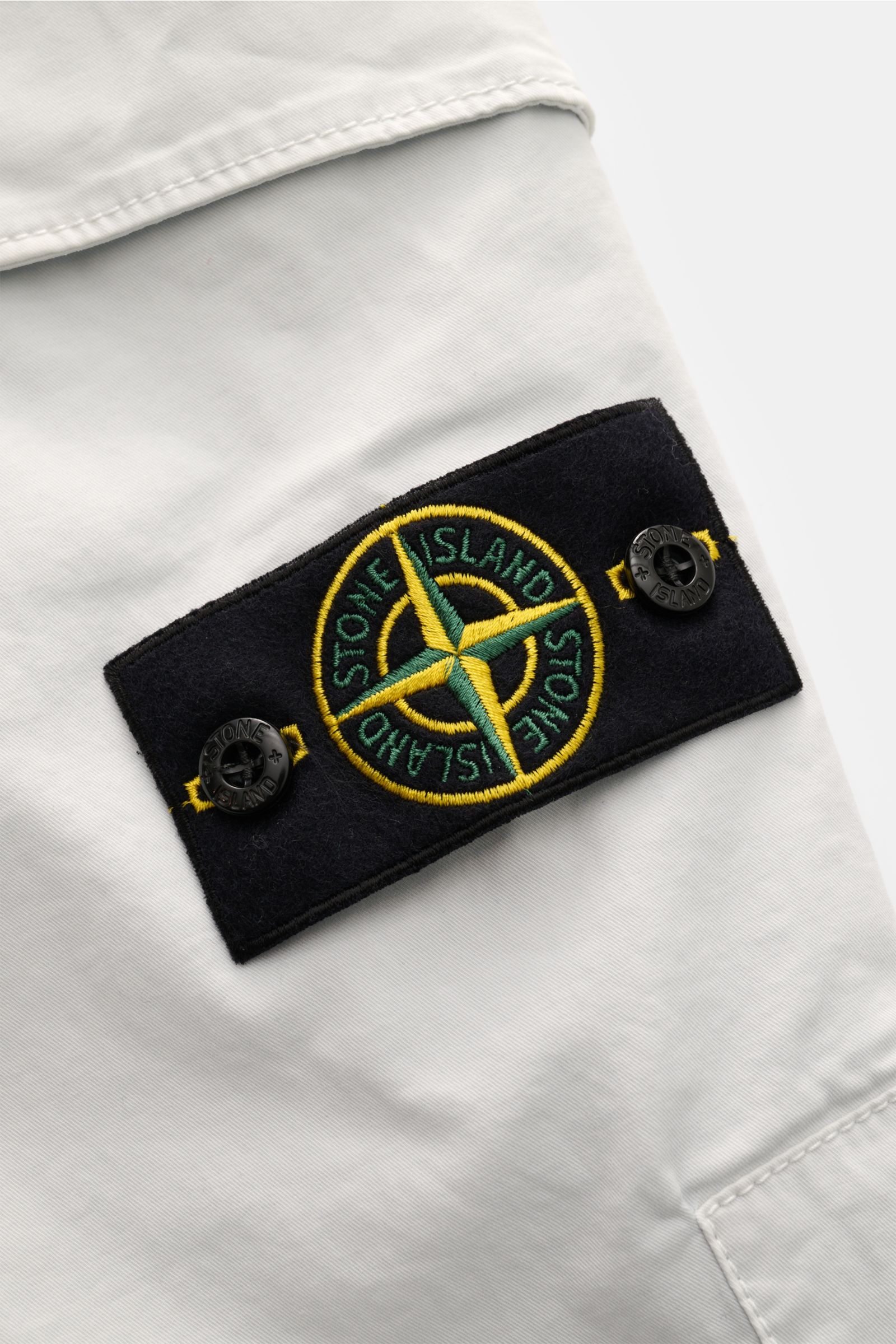Nahaufnahme der Stone Island Cargohose hellgrau aus Baumwoll-Twill mit elastischer Qualität, weichem Griff und typischem Kompass-Badge, frontal fotografiert.
