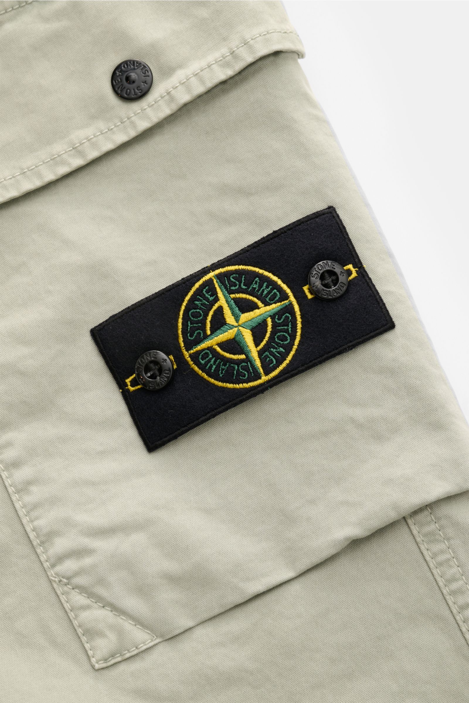 Nahaufnahme der Stone Island Cargohose salbei, Fokus auf das typische Kompass-Badge auf der aufgesetzten Cargotasche aus elastischer, weicher Baumwolle.