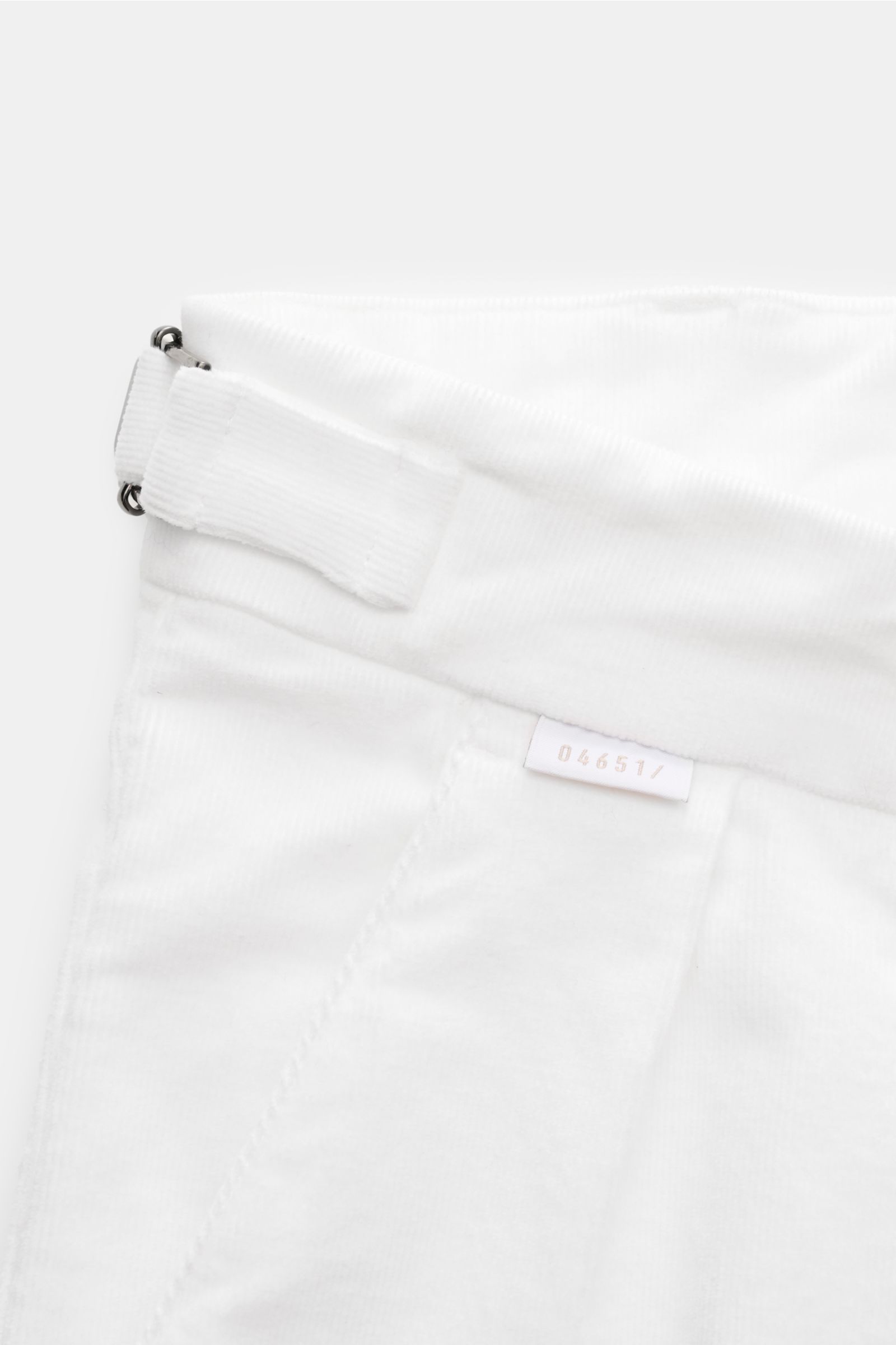 Close-up top-down view of 04651/ A trip in a bag Corduroy trousers off-white, showing cotton corduroy fabric, double pleat, side adjuster, small woven label, soft tapered fit, and fixed hem turn-up details. Retroesque inspiration from 04651/ A TRIP IN A B