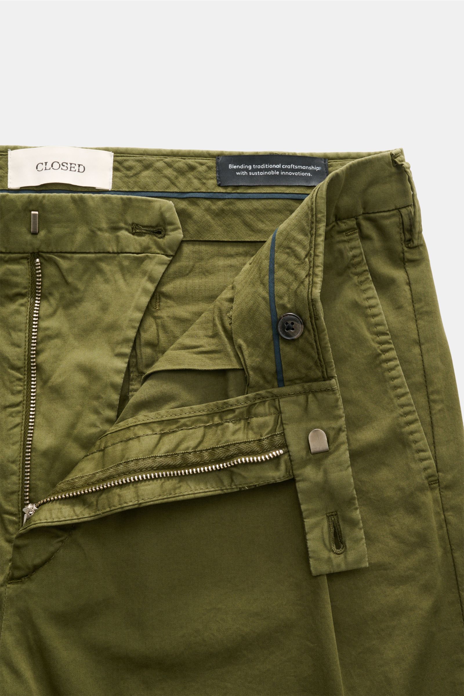 Nahaufnahme der geöffneten Closed Baumwollhose 'Bergen Tapered' oliv von oben, mit sichtbarem Reißverschluss, Bundhaken, Knopf und Label.