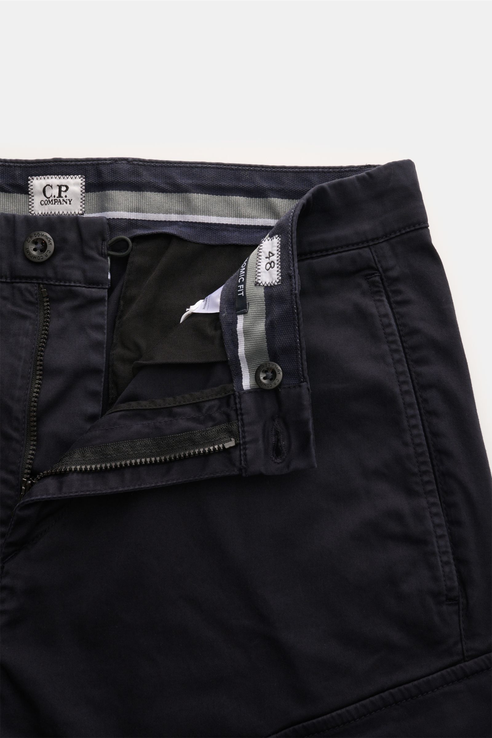 C.P. Company Cargohose dark navy shown from a top-front close-up, highlighting gabardine cotton, stretch, regular fit, zipper, button, side and cargo pockets, and label detail. Urban Utility: Die Cargohose von C.P. COMPANY kombiniert funktionale Details m