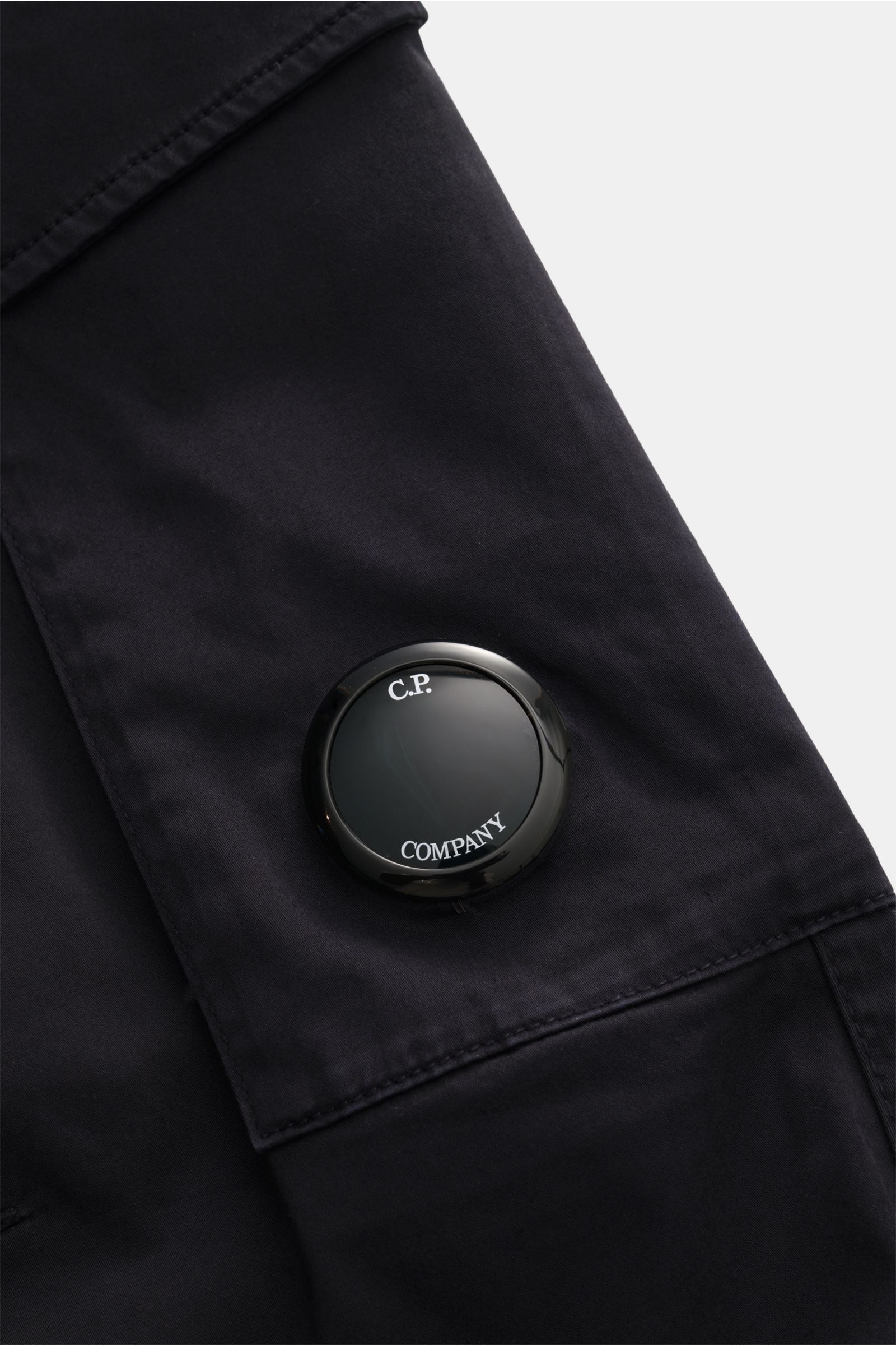 Close-up front view of C.P. Company Cargohose dark navy pocket with label lens detail on gabardine cotton fabric. Urban Utility: Die Cargohose von C.P. COMPANY kombiniert funktionale Details mit entspanntem Fit und hohem Komfort. Gabardine aus Baumwolle, 