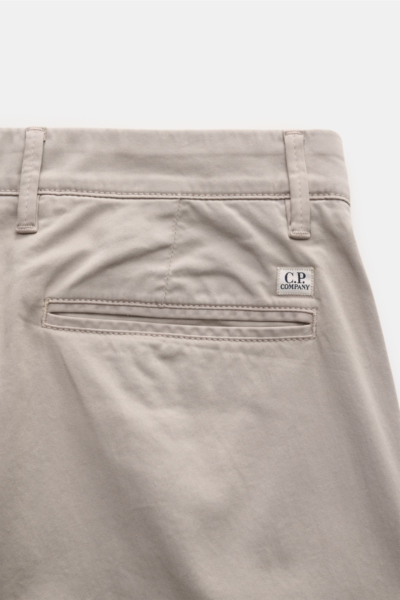 Close-up of the back pocket area of the C.P. Company Cargohose hellgrau, photographed from above, showing soft light grey cotton gabardine fabric, label with lens detail, narrow-cut regular fit, welted back pocket, belt loops, and fine stitching.

Urba