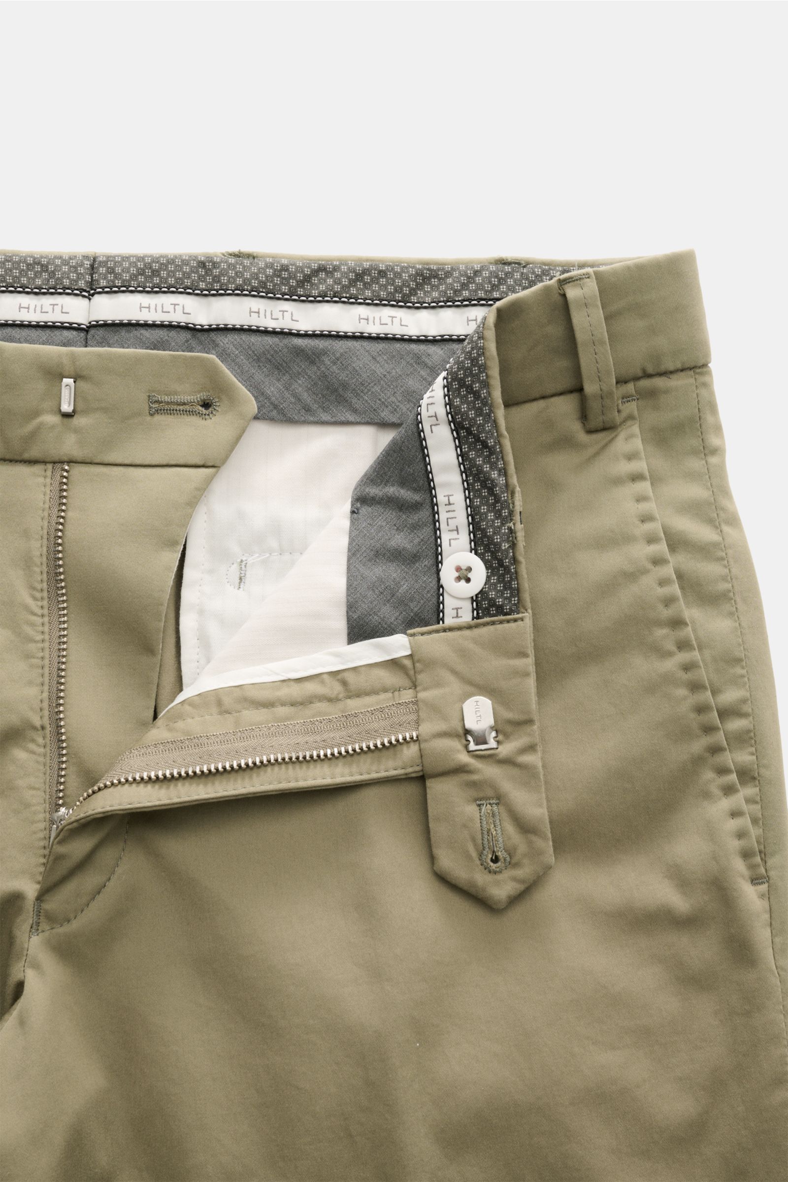 Close-up front view of Hiltl Baumwollhose 'Peaker' oliv showing zipper, slide button, and inner waistband with Hiltl branding, made of lightweight cotton poplin with stretch. Die leichte Hose für jeden Tag: HILTL kreiert mit dem Modell 'Peaker' im Regular