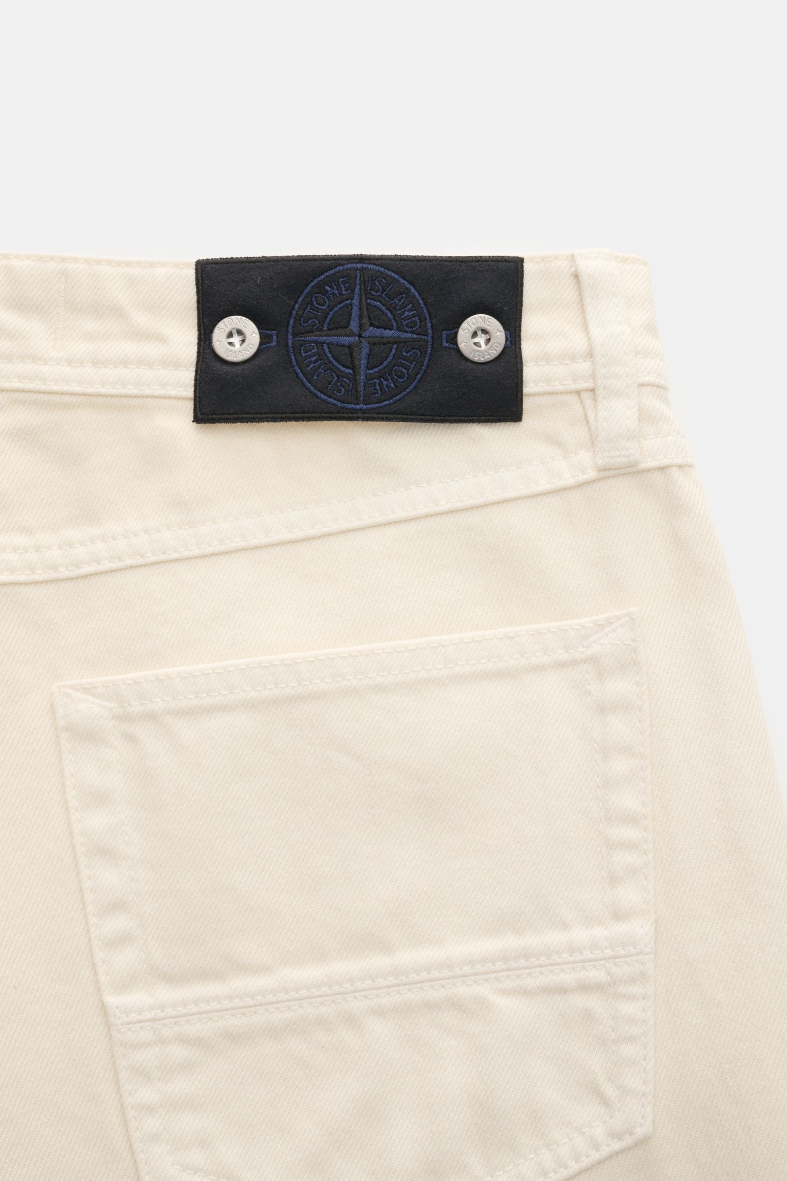 Detailaufnahme der Stone Island Jeans 'Light Bull Denim' creme von hinten mit typischem Kompass-Badge am Bund aus Baumwoll-Denim.