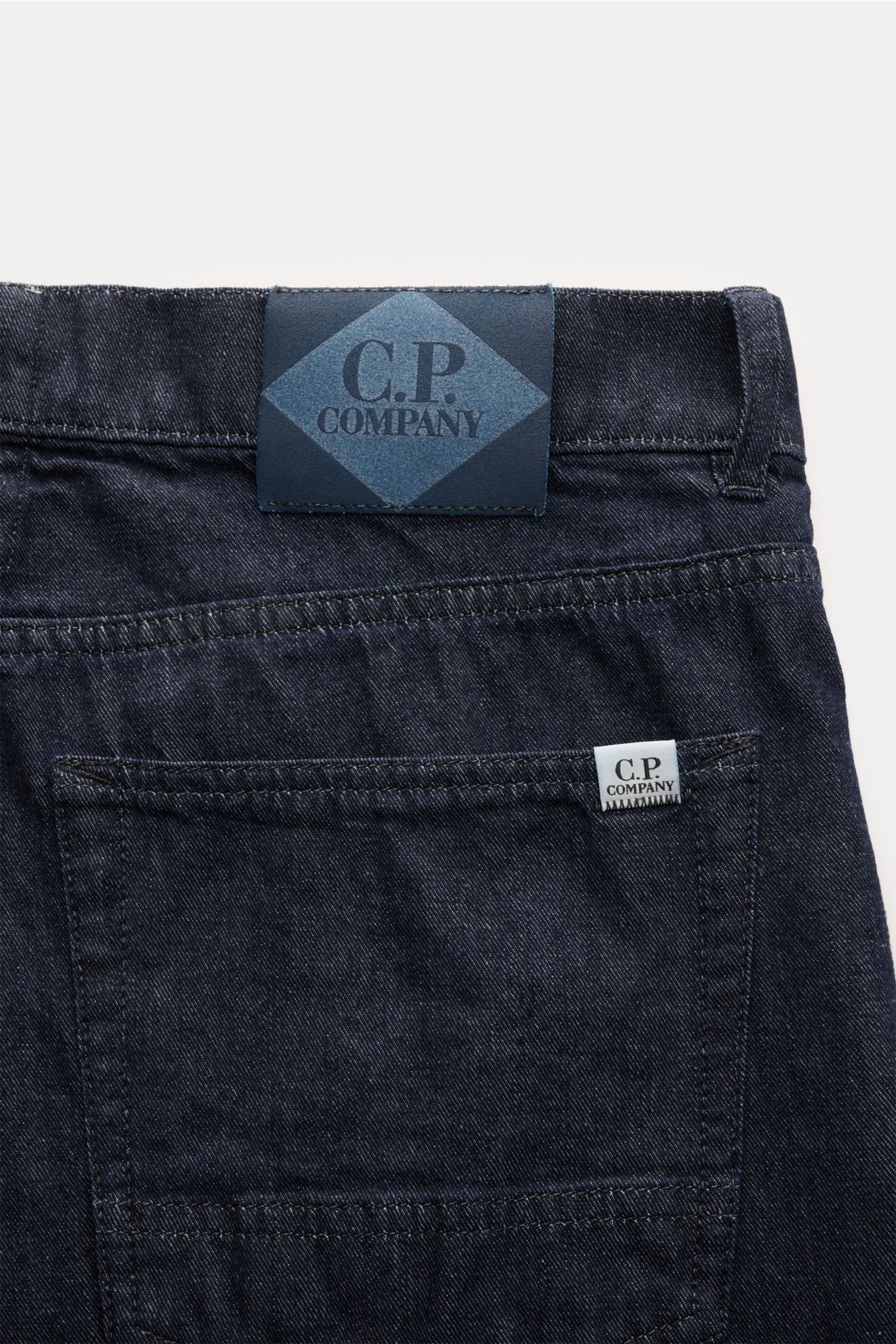 Close-up rear view of C.P. Company Jeans navy showing the waistband with leather patch, back pocket with logo tag, and dark navy denim fabric.