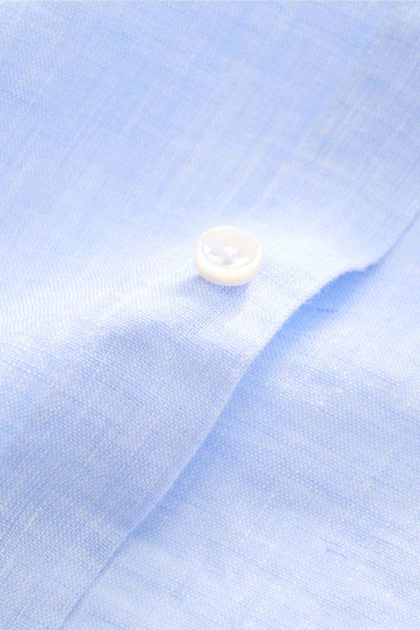 Close-up of the light blue melange fabric and white button of the von Braun Leinenhemd Haifisch-Kragen hellblau meliert, photographed from above, showing the linen’s texture and fine stitching.

Mühelose 