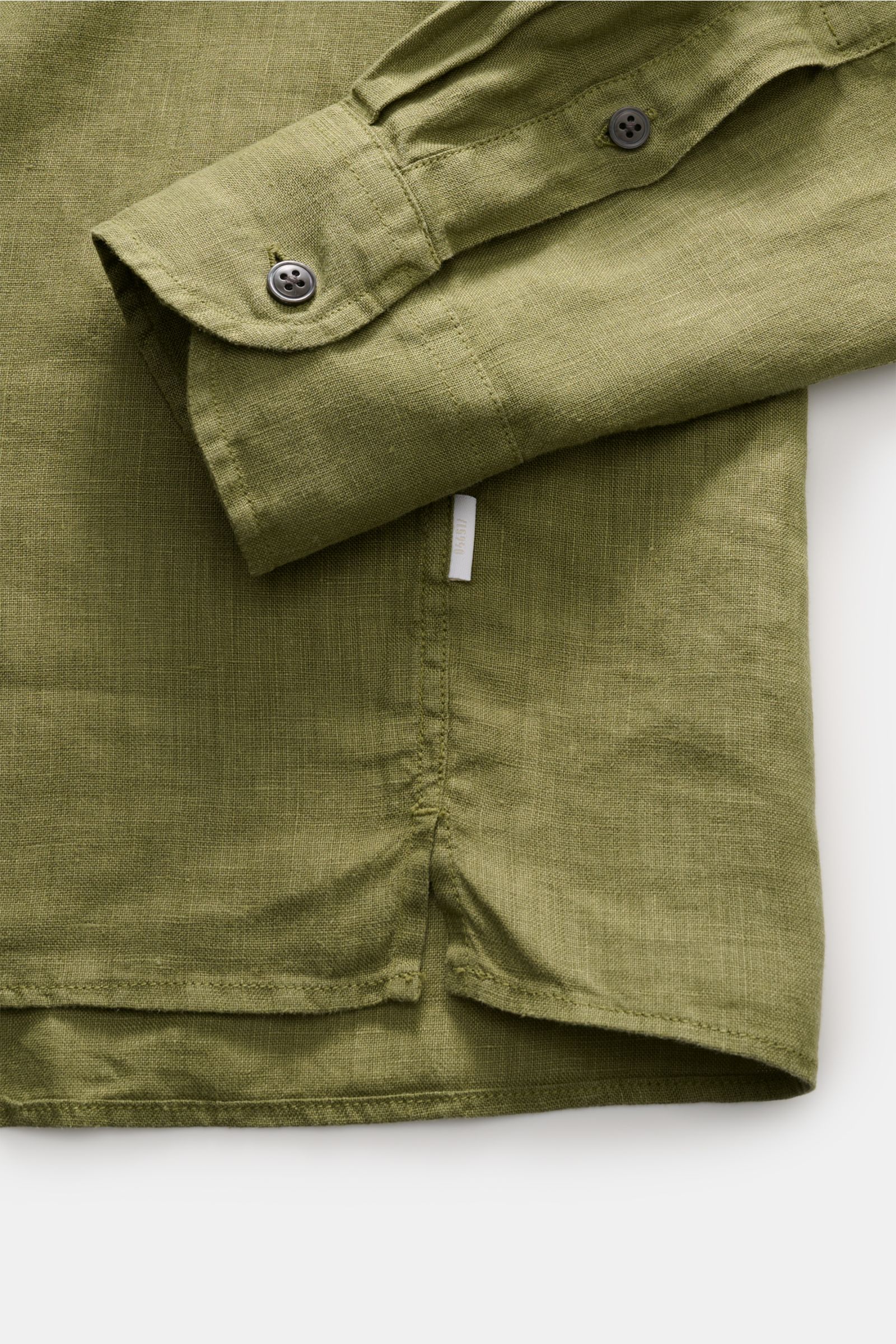 Close-up side view of the olive green 04651/ A trip in a bag Leinenhemd Kent-Kragen oliv showing sleeve cuff, side seam with small white label, and hem with slit, made of pure linen with light, airy texture.

Mühelose 