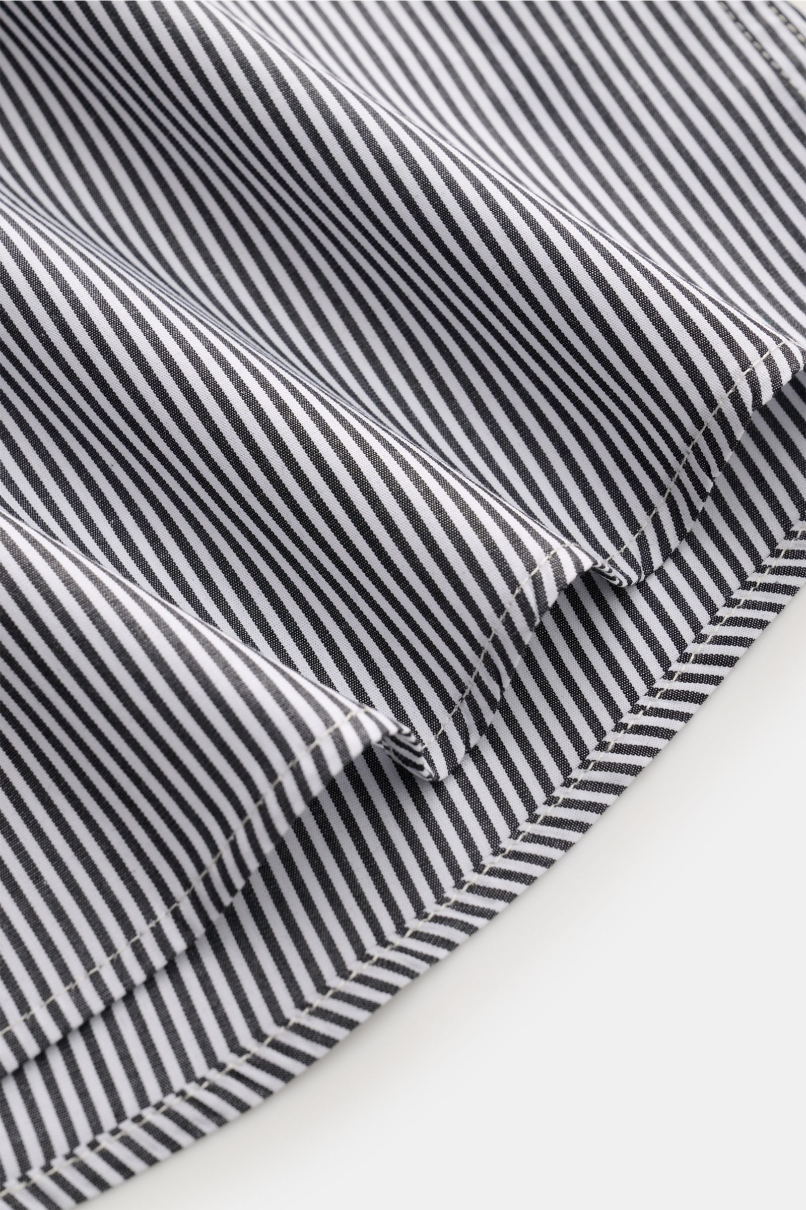 Close-up of the hem of the A.P.C. Casual Hemd Kent-Kragen schwarz/weiß gestreift, showing fine black and white stripes, smooth pure cotton fabric, and precise stitching from a side angle, highlighting its slightly rounded hem. Klassischer Look, modern int