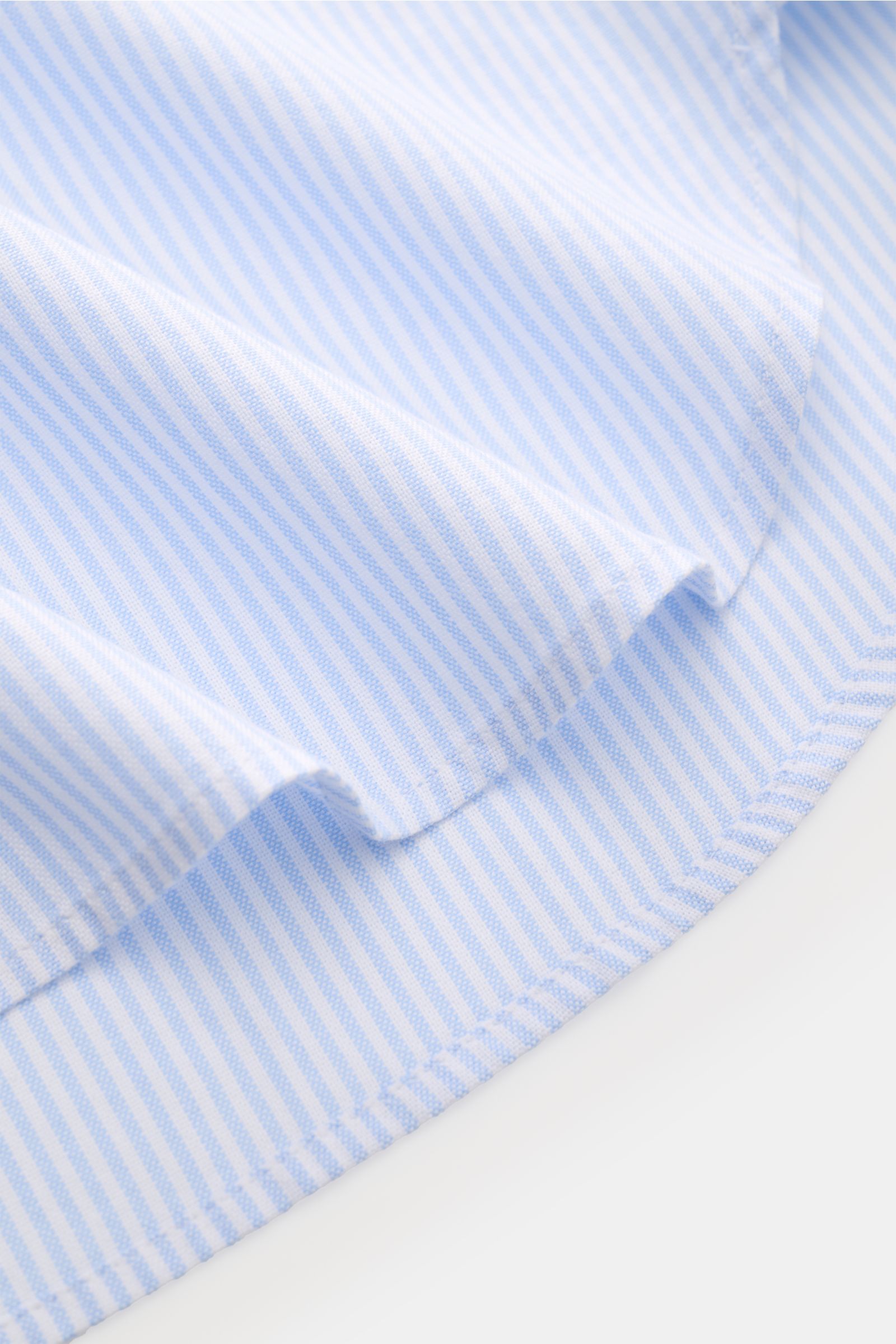 Close-up of the hem and fabric texture of the Officine Générale Oxford-Hemd Button-Down-Kragen weiß/hellblau gestreift, showing soft white and light blue stripes from a side angle, highlighting the finely woven pure cotton Oxford material.

Zeitloses E