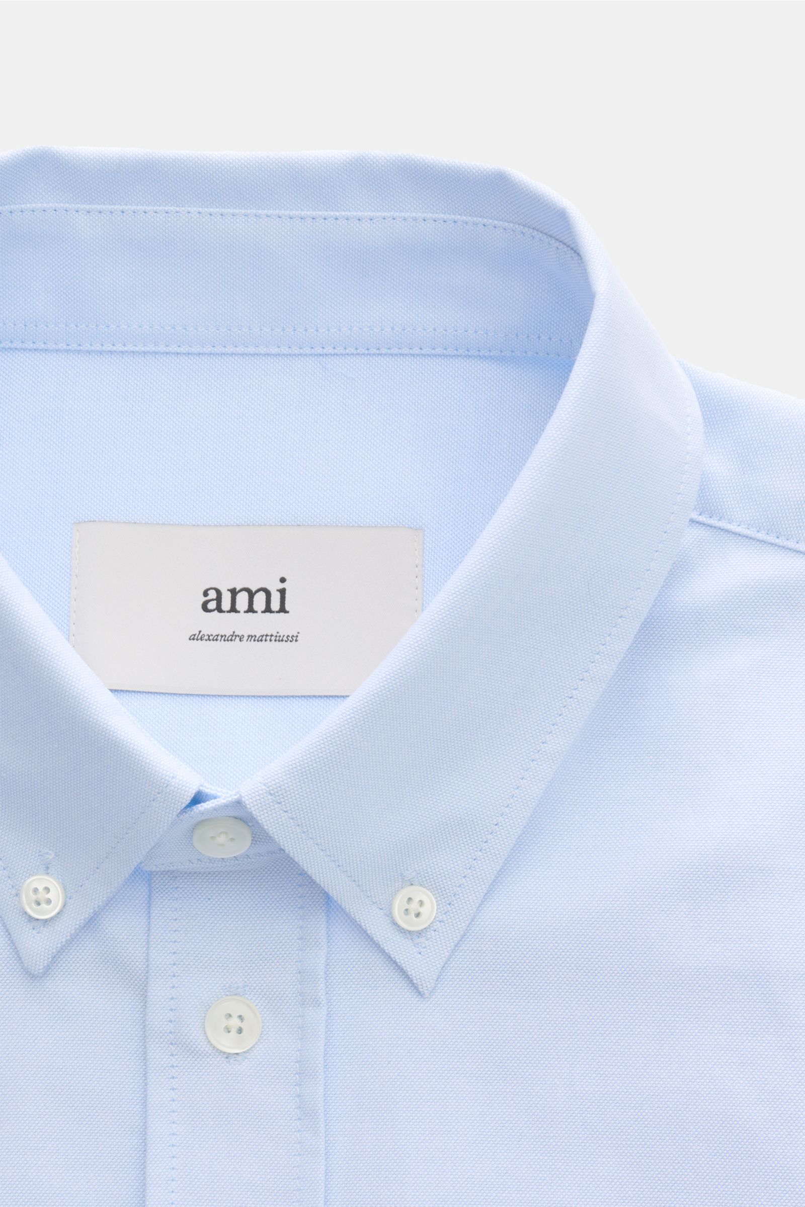 A close-up front view of an Ami Paris Oxford shirt button-down collar light blue, showing the button-down collar, patch button placket with mother-of-pearl buttons, and signature heart stitching. For relaxed casual styles and more formal looks: This elega