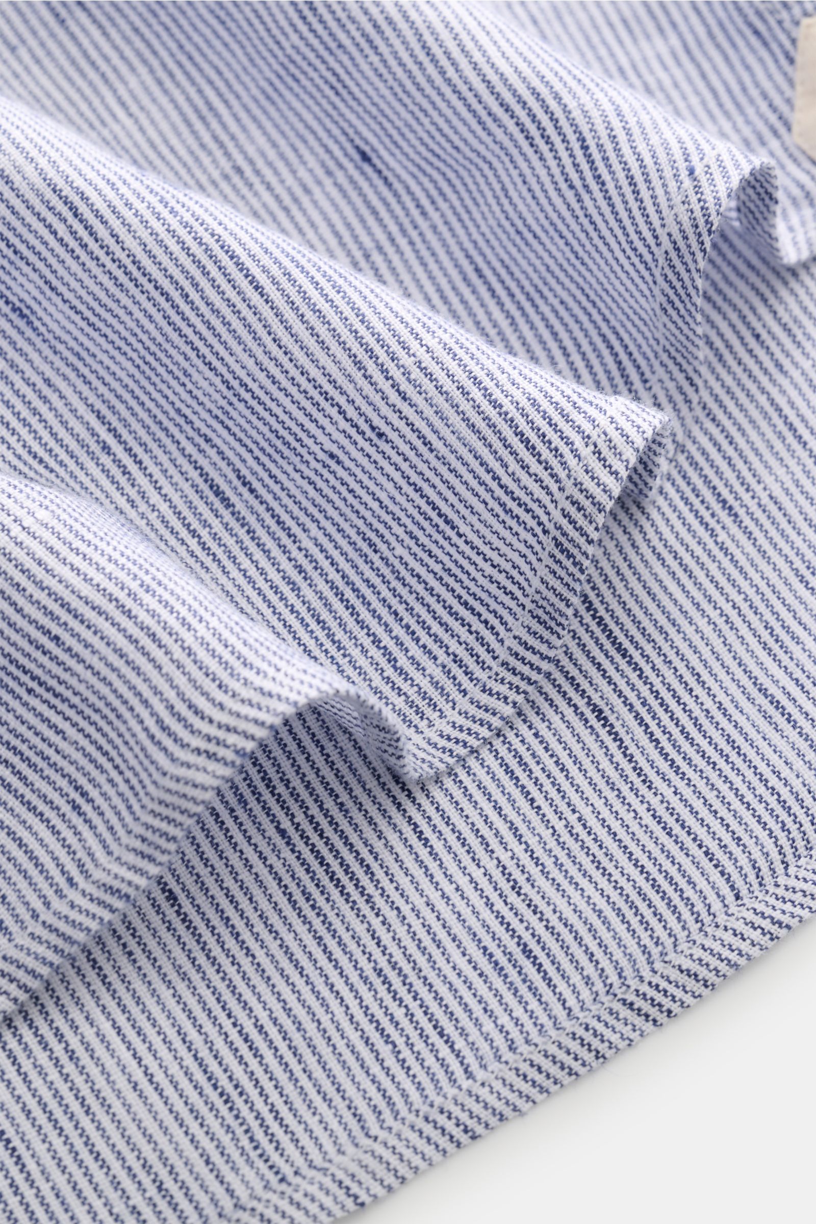 Nahaufnahme des von Braun Leinenhemd Haifisch-Kragen navy/weiß gestreift, Detailansicht der feinen Streifen und Struktur aus reinem Leinen, luftig und weich, gerundeter Saum sichtbar, fotografiert aus seitlicher Perspektive.