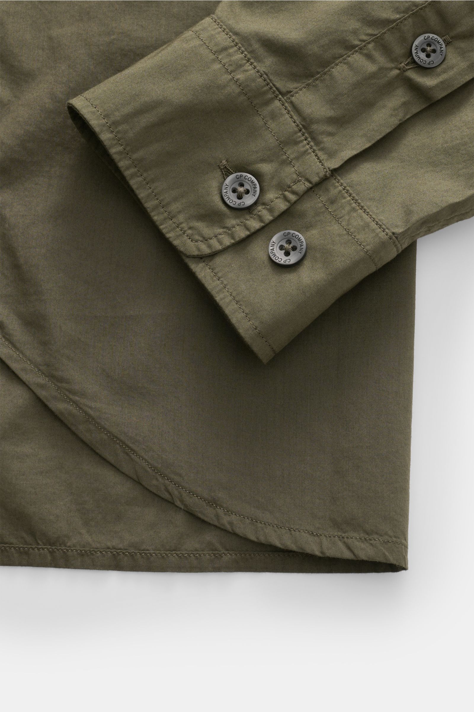 Nahaufnahme des Ärmels und Saumverlaufs des C.P. Company Casual Hemd schmaler Kragen dark olive aus reiner Baumwolle, leicht und garment dyed, mit Logo-Knöpfen.