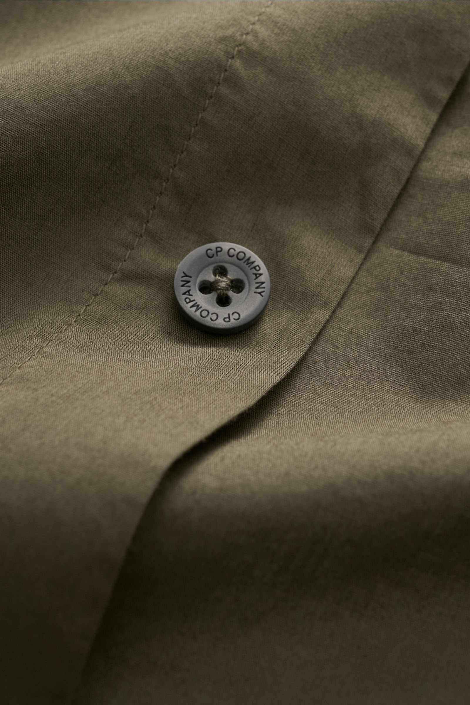 Nahaufnahme eines dunkelolivfarbenen C.P. Company Casual Hemd schmaler Kragen dark olive aus reiner Baumwolle, Fokus auf Knopf mit Markenprägung, weicher Griff, garment dyed Stoff, Detailansicht von schräger Perspektive.