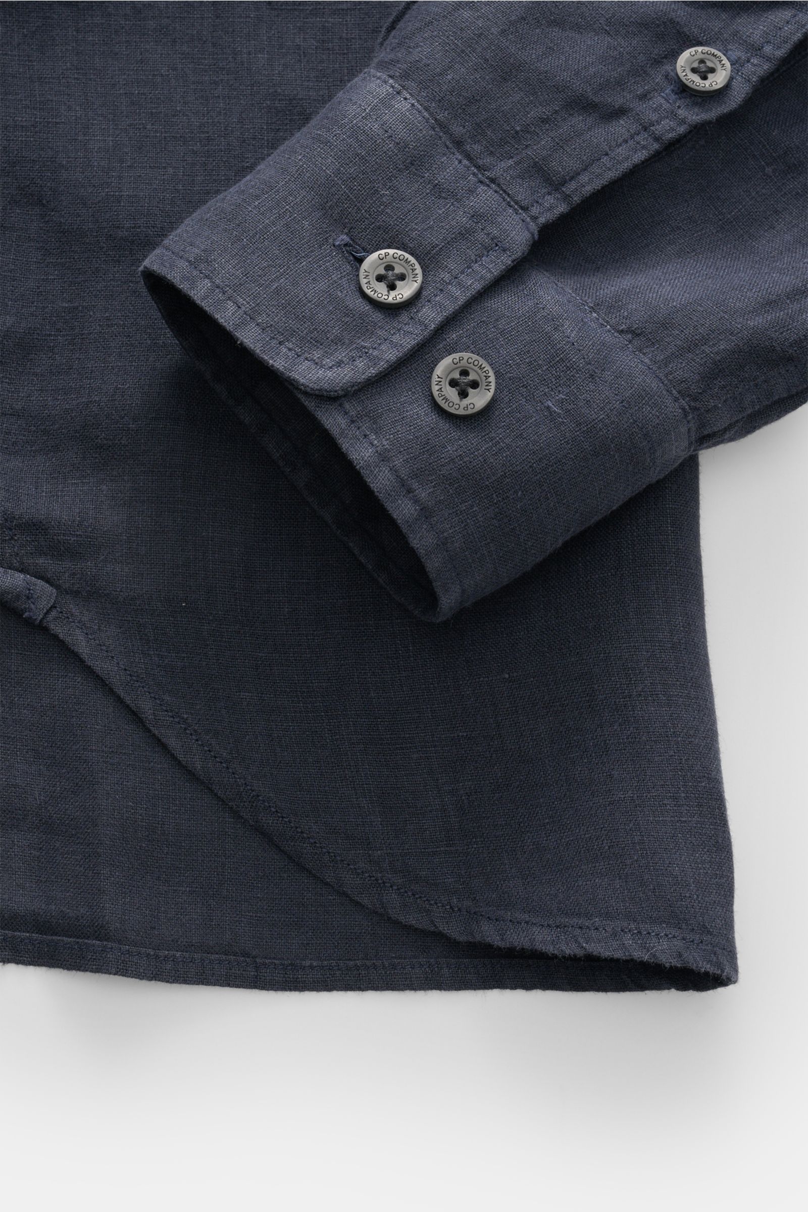C.P. Company Linen shirt slim collar navy shown from a close-up top-down perspective, highlighting pure linen fabric, soft feel, garment-dyed navy color, classic barrel cuffs with logo buttons, and rounded hem with slightly elongated back. 
