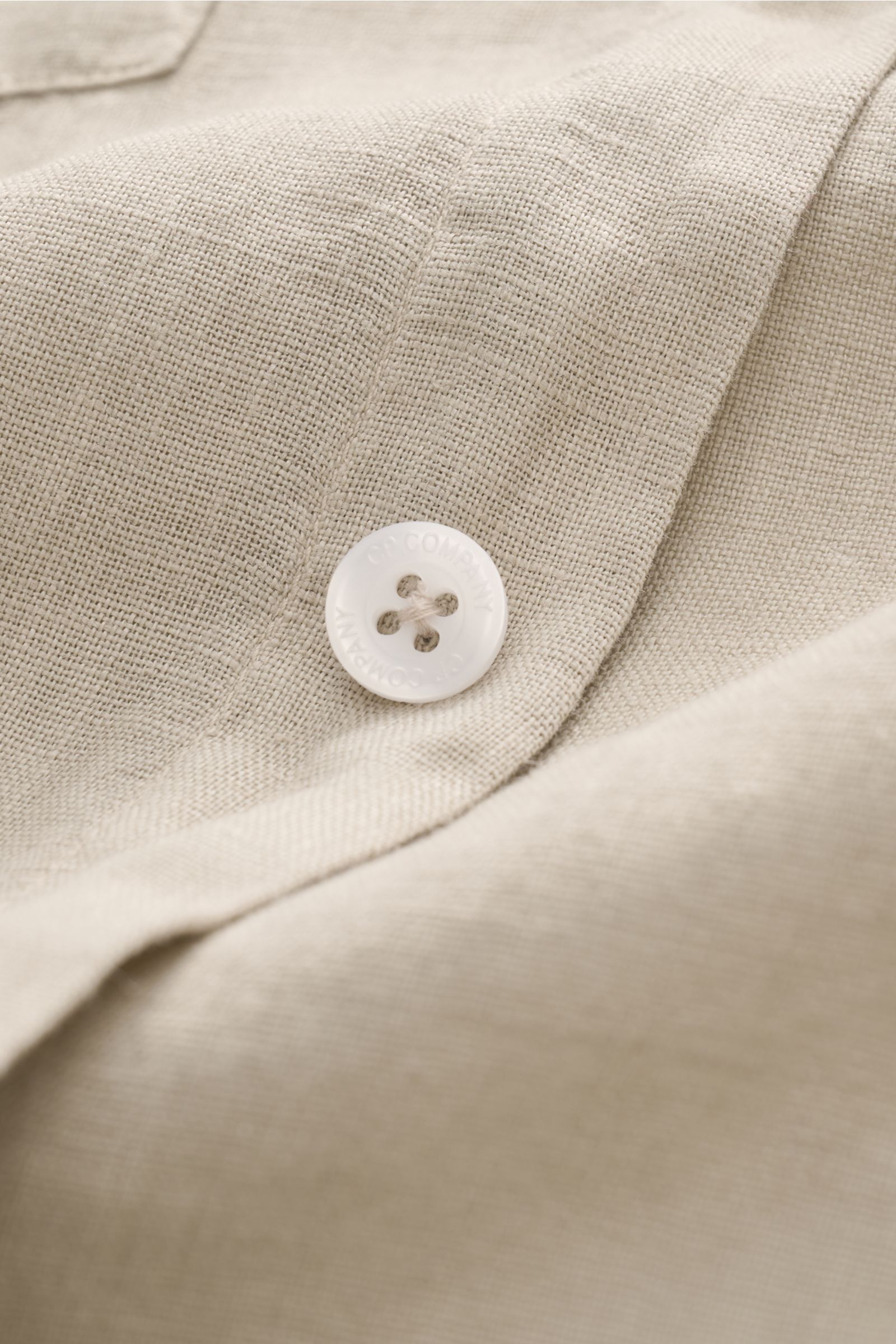 Close-up front view of C.P. Company Linen shirt narrow collar sand, showing pure linen fabric texture, white branded button, and stitching details. 