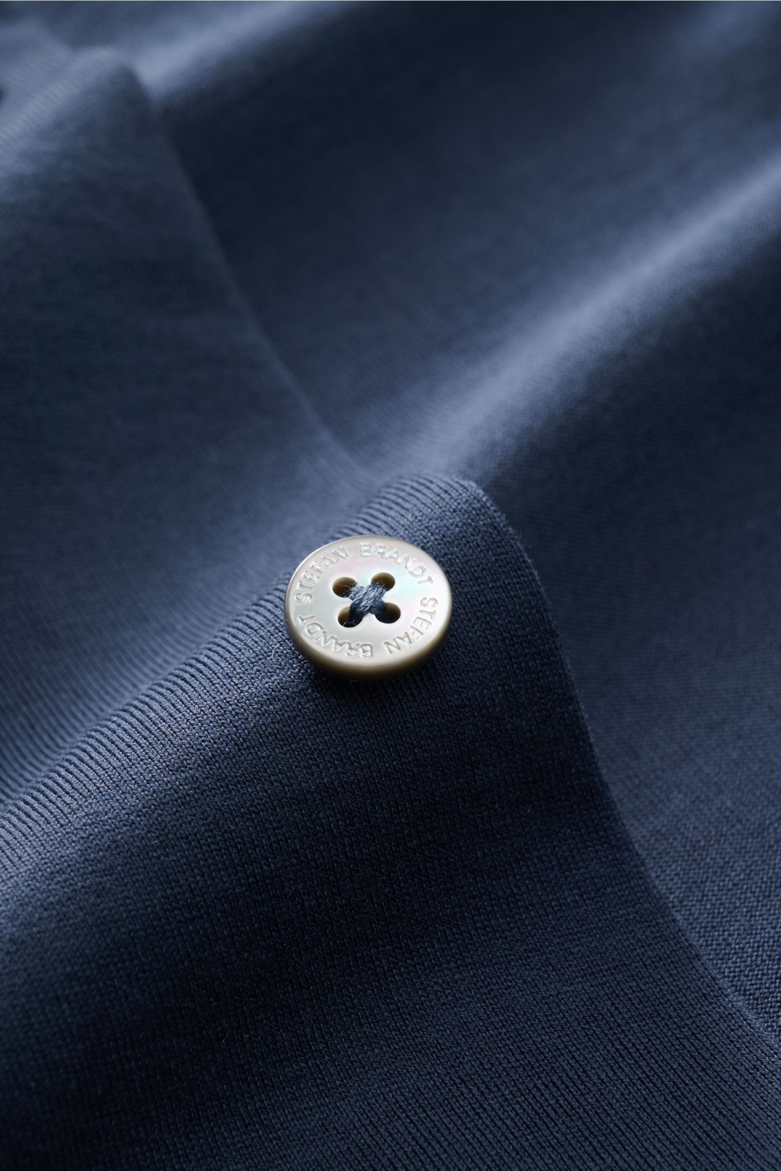 Close-up top-down view of a pearl button on the dark blue fabric of the Stefan Brandt Jersey-Hemd 'Otis' Haifisch-Kragen dunkelblau.

Description: Flexibler, geschmeidiger Jersey aus reiner Pima-Baumwolle lässt das cleane Casual Hemd 'Otis' von STEFAN 