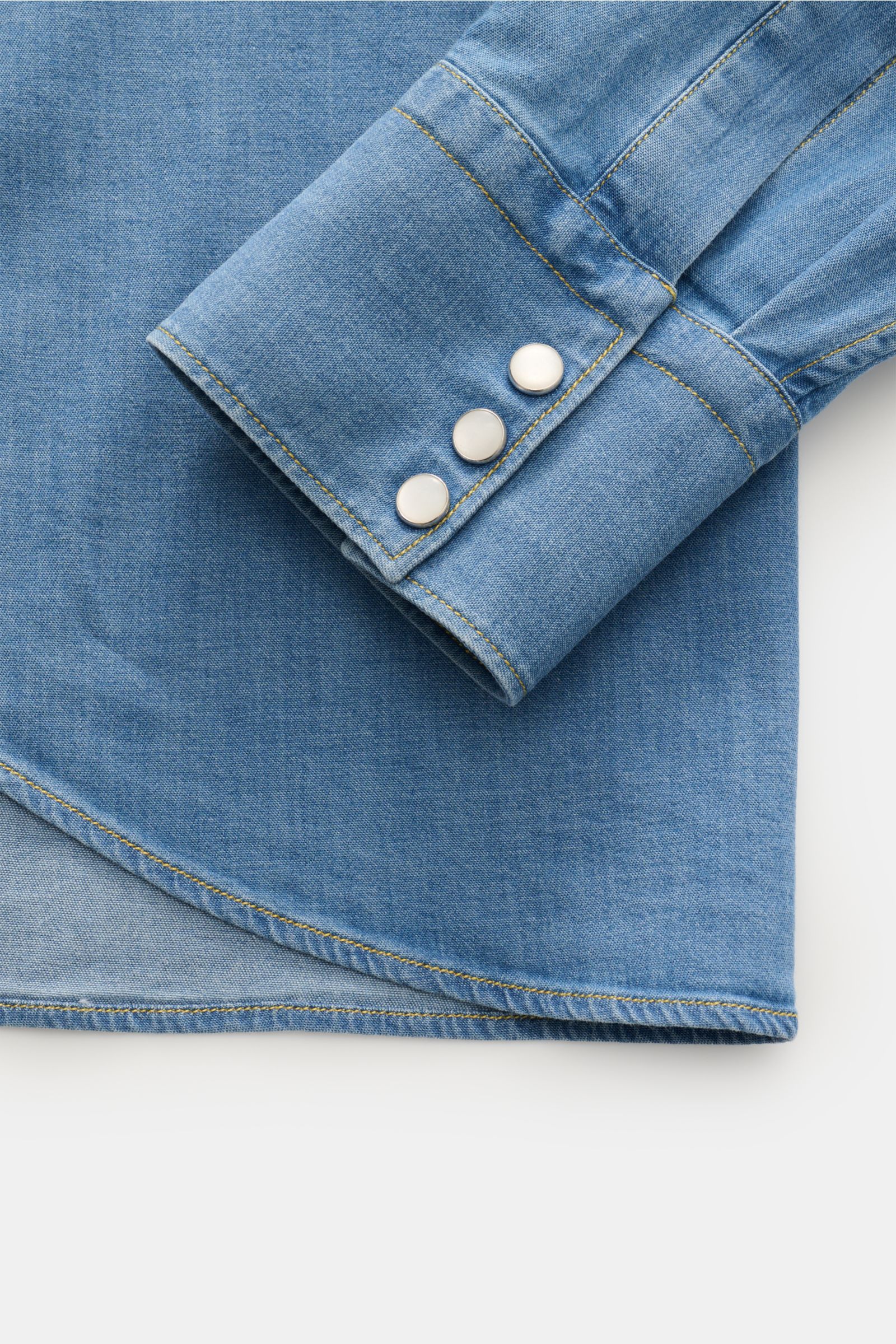 Close-up of the sleeve and rounded hem of the Gherardi Chambray shirt Kent collar smoky blue, showing soft stretchy cotton chambray fabric, western-style shoulder yoke, patch snap button placket, and barrel cuffs with snap buttons, photographed from above