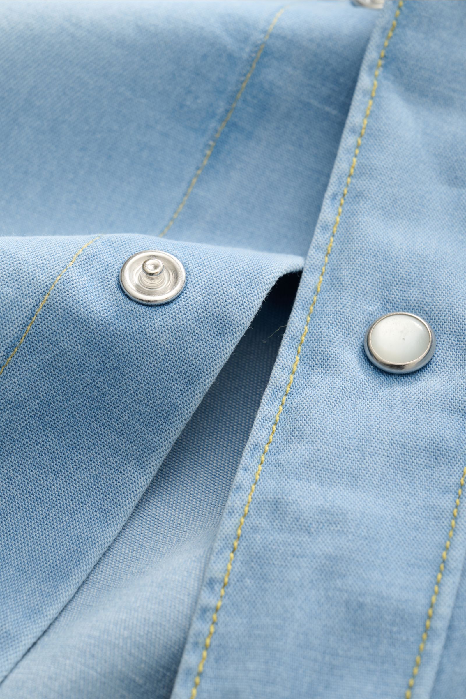 Close-up of Gherardi Chambray-Hemd Kent-Kragen hellblau showing light blue chambray fabric with silver snap buttons and yellow stitching, photographed from a top-down perspective.

Description: Legeres Chambray-Hemd vom Experten GHERARDI: Dieses Modell