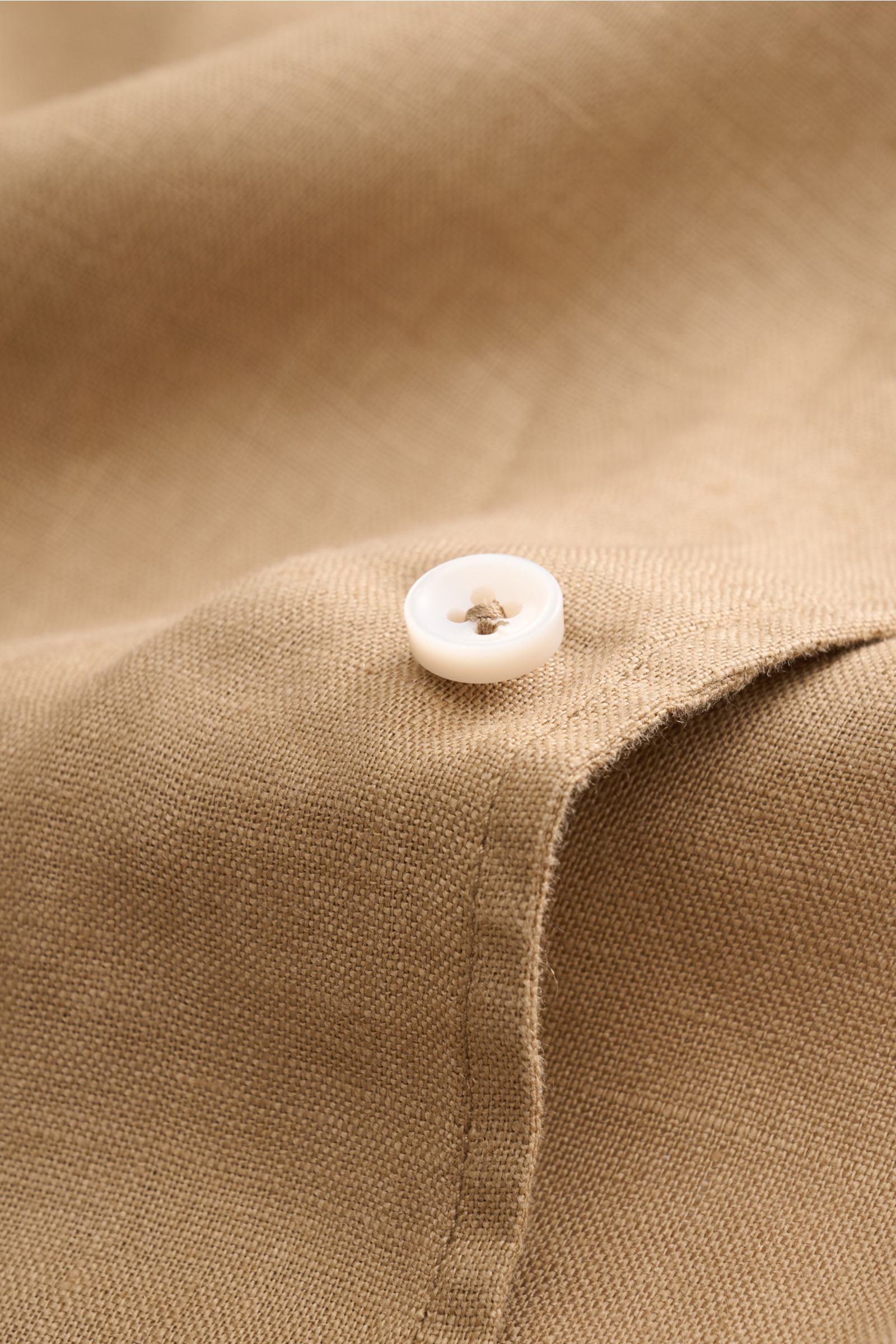 Aurélien Leinenhemd Haifisch-Kragen sand close-up from above showing sand-colored pure linen fabric with a white button, soft texture, slim fit. Ein wenig Urlaubsfeeling gefällig? Das vielseitig einsetzbare Hemd der Marke AURÉLIEN wird mit seinem feine