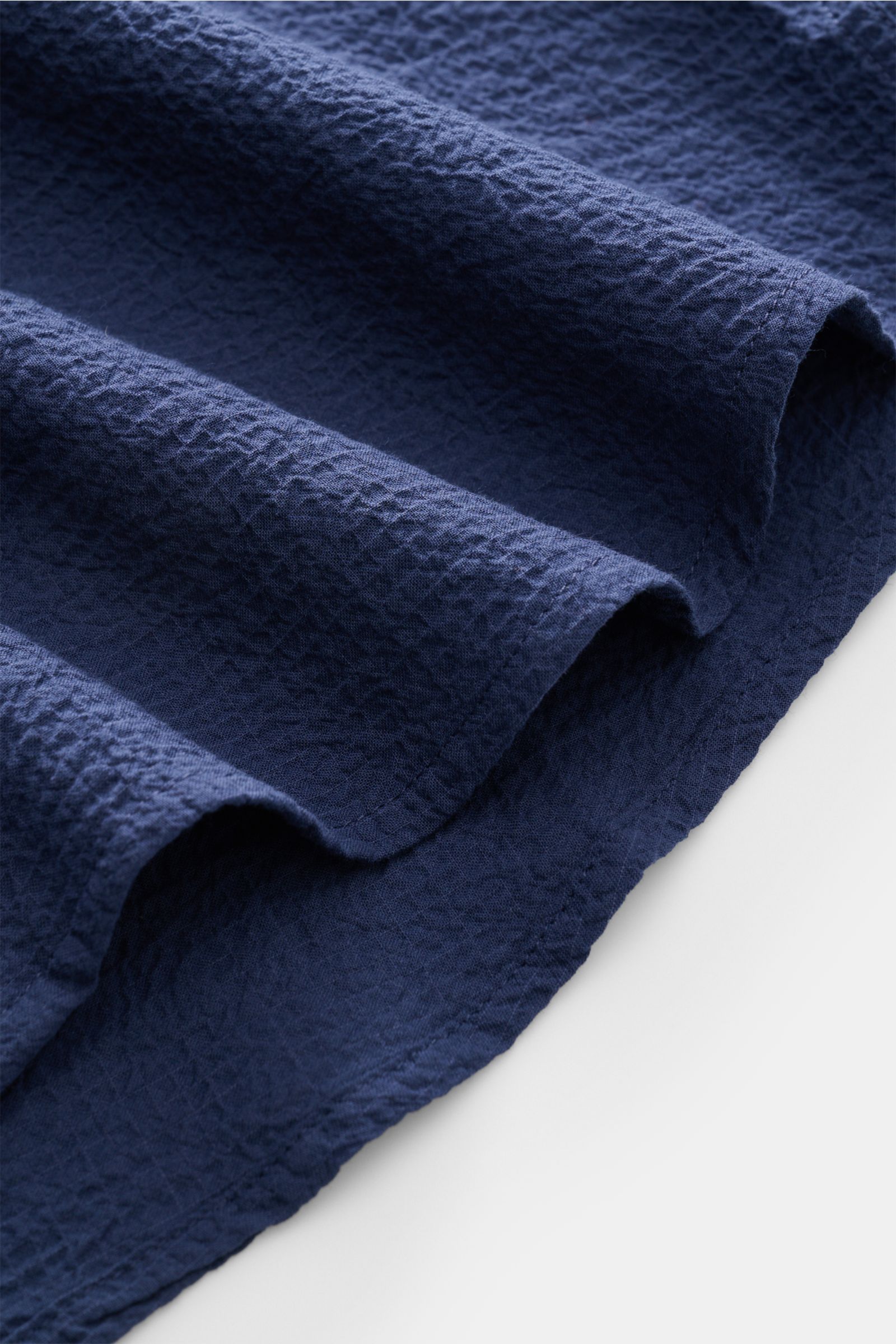 Close-up of the dark blue Aurélien Seersucker-Hemd Haifisch-Kragen fabric showing its lightweight, textured cotton seersucker material with slight elasticity, photographed from a side angle highlighting the slightly rounded hem edge. 

Sommerlicher Eye
