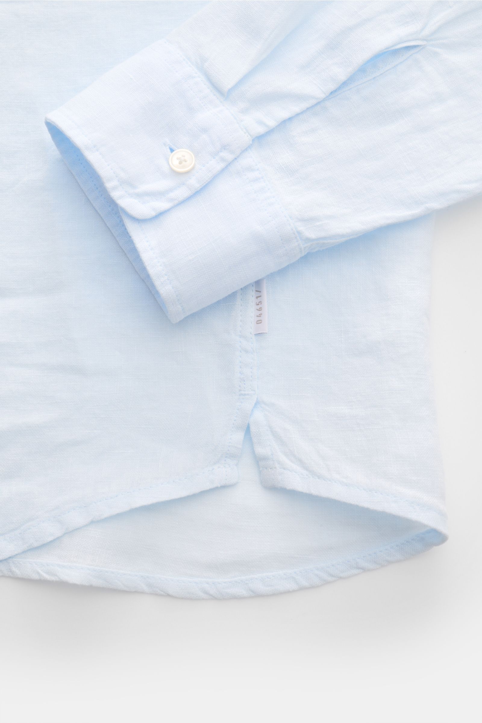 Close-up front-bottom view of 04651/ A trip in a bag Leinenhemd schmaler Kragen pastellblau showing light blue pure linen fabric, narrow collar, French placket, classic cuff with button, small side seam label, rounded hem with short side slits. 