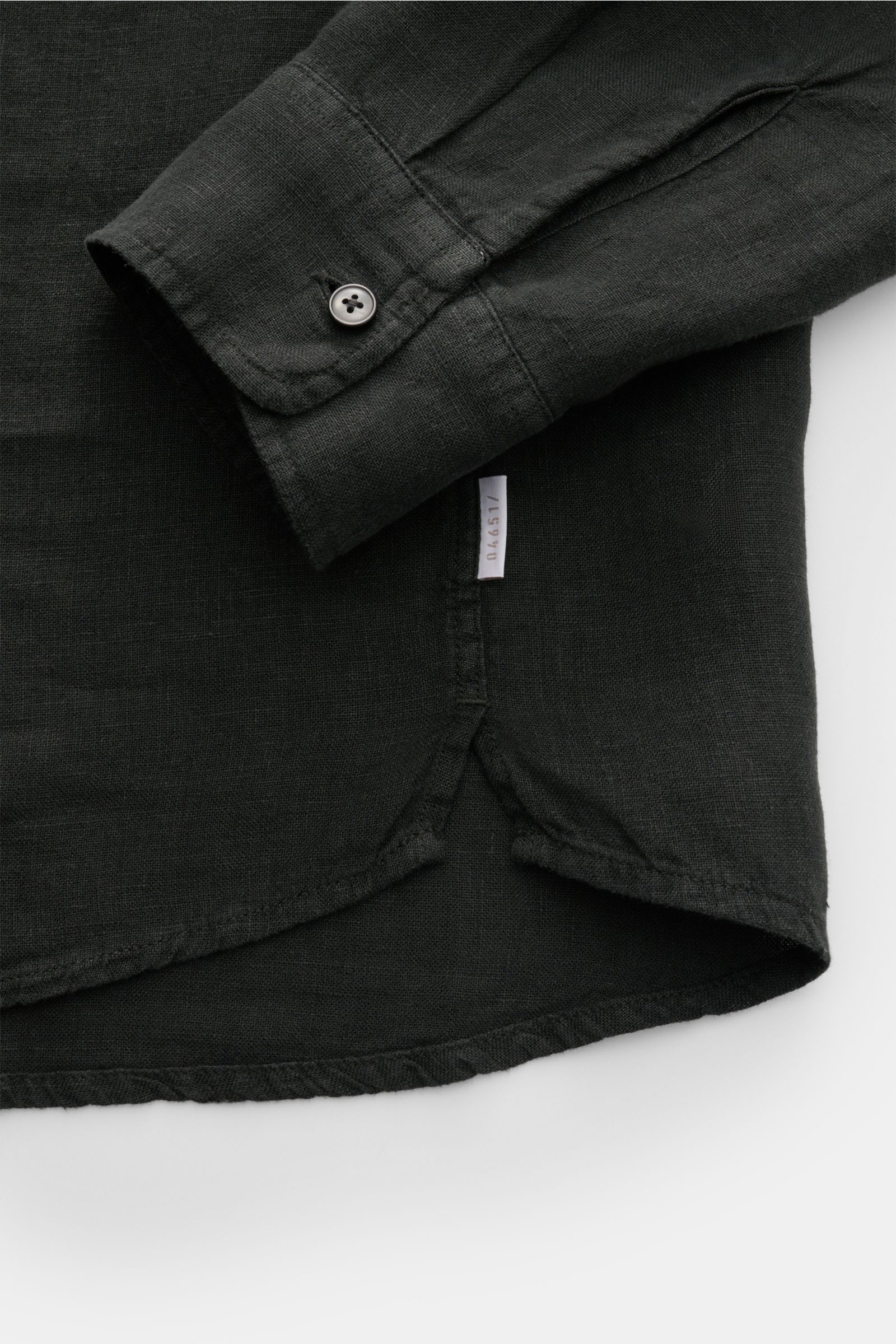 Black linen shirt cuff and lower side hem with small white label visible, photographed from a close-up top-down angle; 04651/ A trip in a bag Leinenhemd schmaler Kragen schwarz. Mühelose 