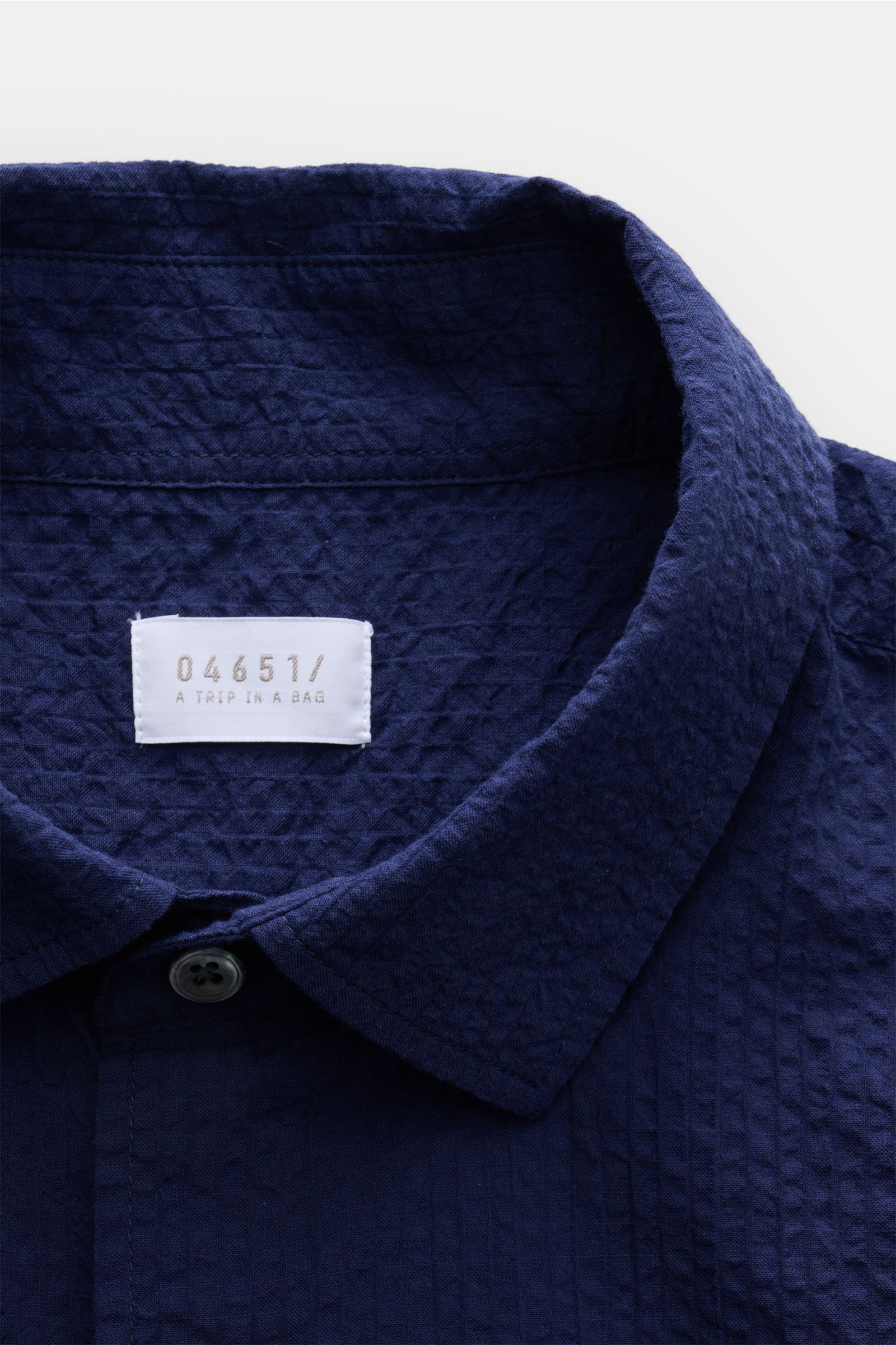 Close-up front view of the navy 04651/ A trip in a bag Seersucker shirt Kent collar navy, showing textured seersucker cotton fabric, Kent collar, single button, and brand label inside collar. Effortlessly casual: This shirt made of pure cotton by 04651/ A