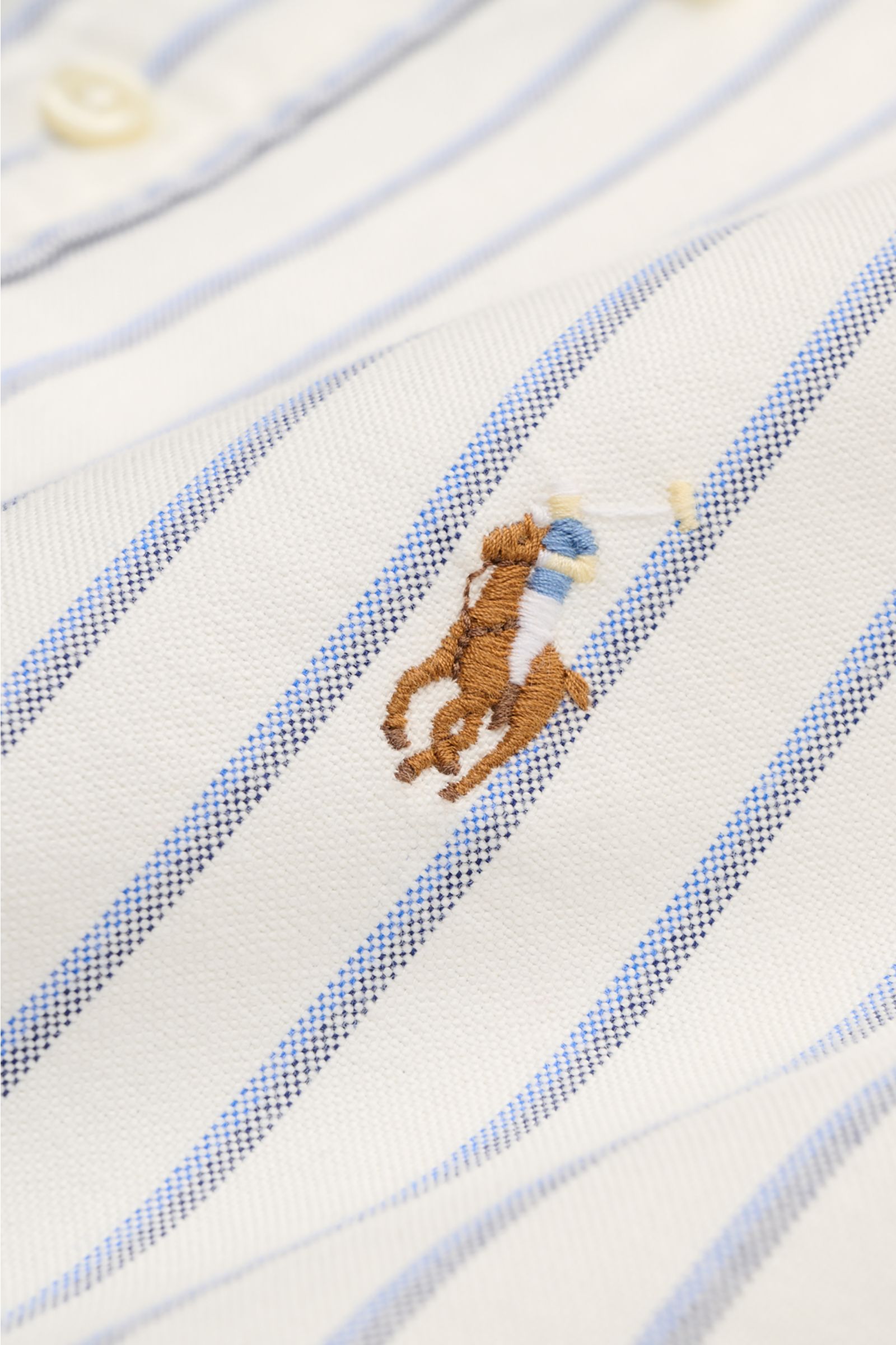 Nahaufnahme des Polo Ralph Lauren Oxford-Hemds Button-Down-Kragen offwhite/blau/schwarz gestreift mit Polospieler-Stitching, weichem Baumwollstoff und gerundeter Saumkante, aufgenommen von schräger Perspektive.