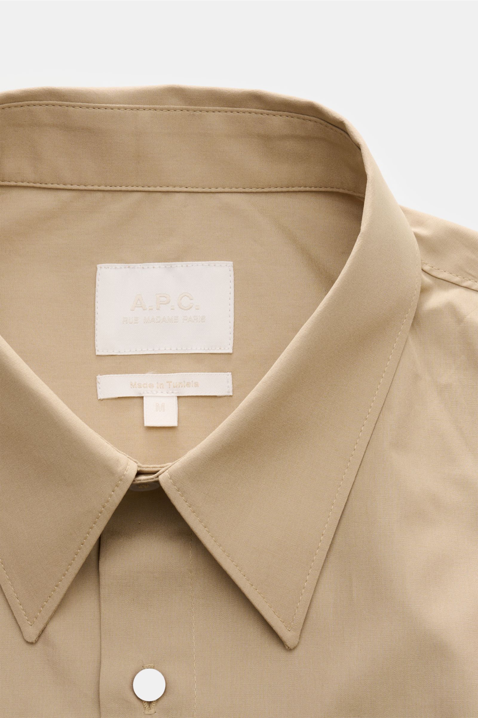 A close-up front view of the A.P.C. Kurzarmhemd Kent-Kragen sand, a sand-colored short-sleeve shirt with a Kent collar, made of pure cotton, featuring a smooth texture, oversized fit, French button placket, patch chest pocket, back yoke with pleat, and a 