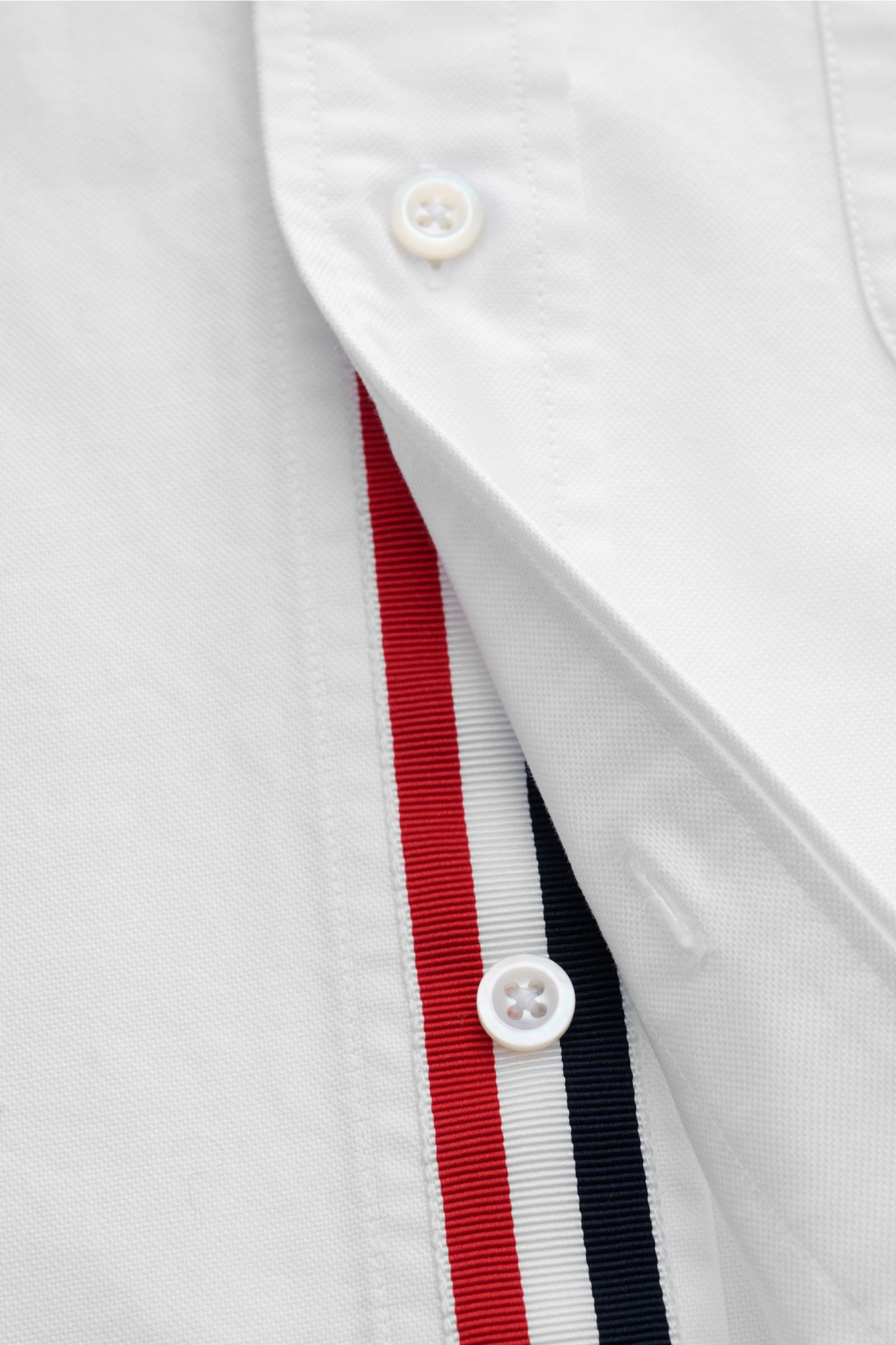 Close-up front view of Thom Browne Short-sleeve shirt button-down collar white showing button placket with red, white, and navy stripes, pure cotton Oxford fabric, soft feel, regular fit, patch breast pocket, back yoke with inverted pleat and hanging loop