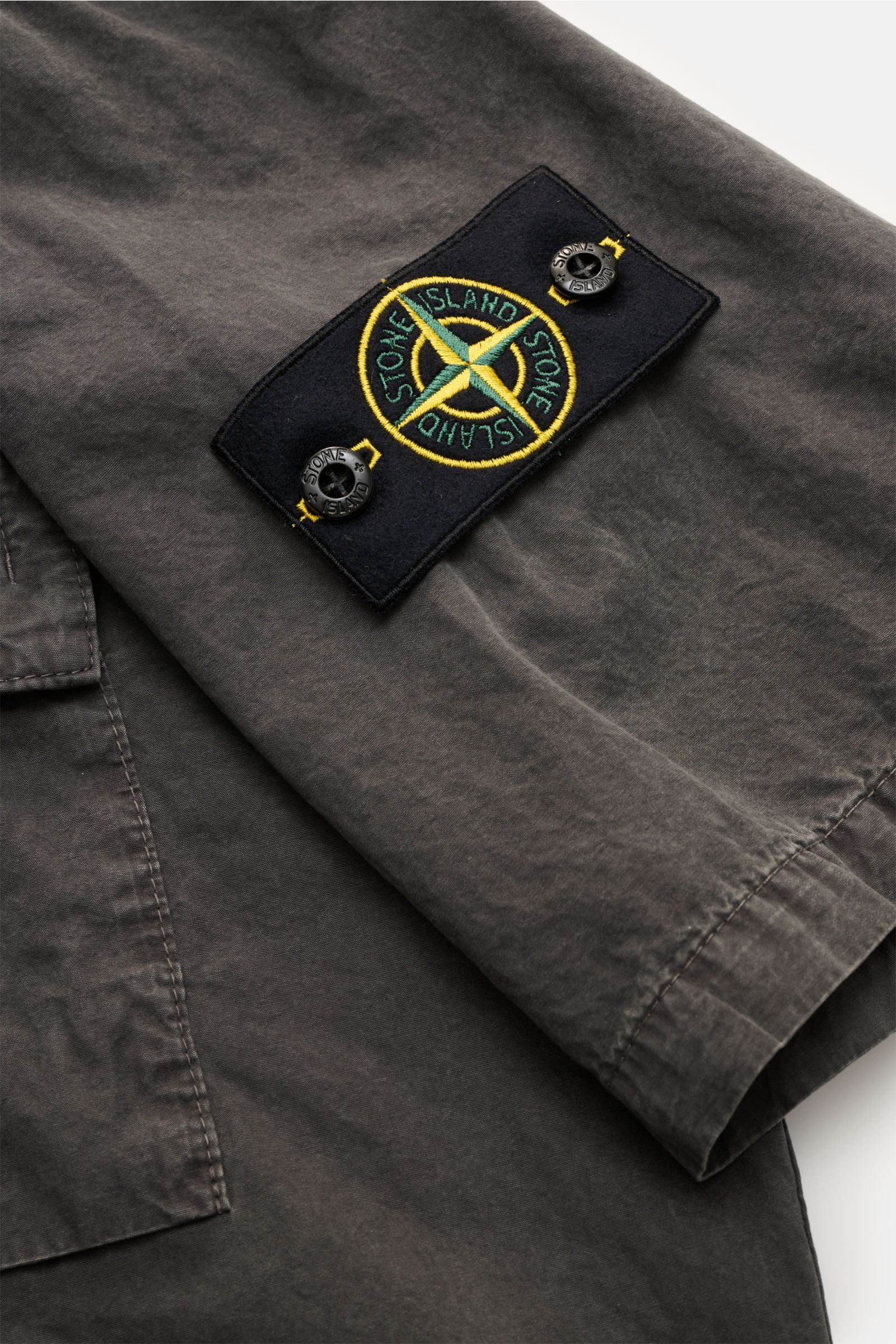 Close-up side view of the sleeve of the Stone Island Short sleeve shirt 'Brushed Canvas' Kent collar anthracite, showing the compass badge on soft, garment-dyed pure organic cotton fabric with a concealed snap button closure, patch breast pocket with butt