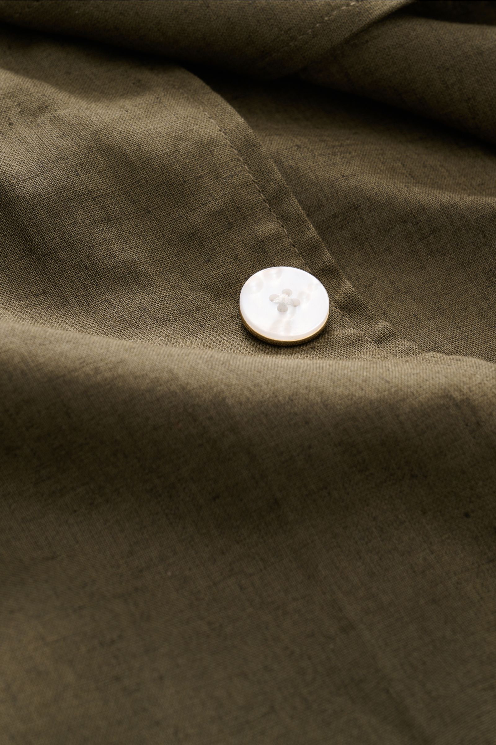 Close-up view of the fabric and white button detail on the Ché Kurzarmhemd 'Inigo' Kubanischer Kragen dark olive, a linen-cotton mix summer shirt with a textured feel, oversized fit, Cuban collar, chest pockets, French button placket, and crochet hem deta