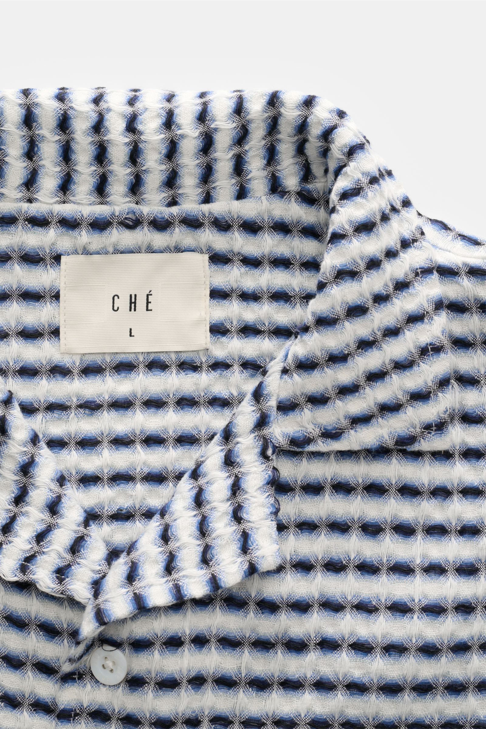 Close-up front view of Ché Leinen-Kurzarmhemd 'Riita' Kubanischer Kragen blau/weiß gemustert showcasing its intricate blue and white woven waffle piqué pattern and the CHÉ label. 

Description: Im Sommer kreiert das Leinen-Kurzarmhemd 'Riita' mit Kuban