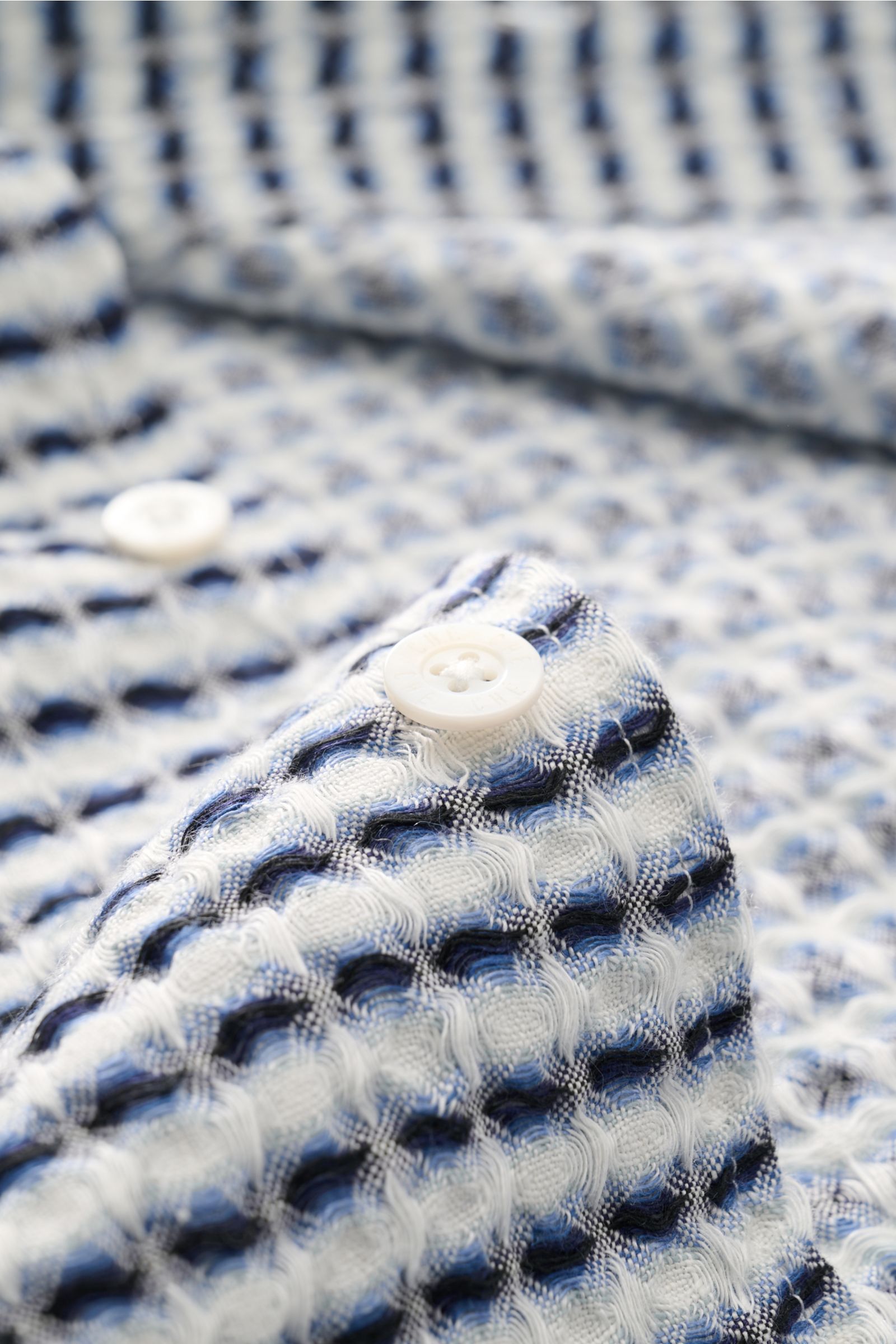 Close-up front view of Ché Leinen-Kurzarmhemd 'Riita' Kubanischer Kragen blau/weiß gemustert showing white pearl-effect buttons and intricate blue and white waffle piqué linen fabric with a coarse, airy knit pattern. Im Sommer kreiert das Leinen-Kurzarmhe