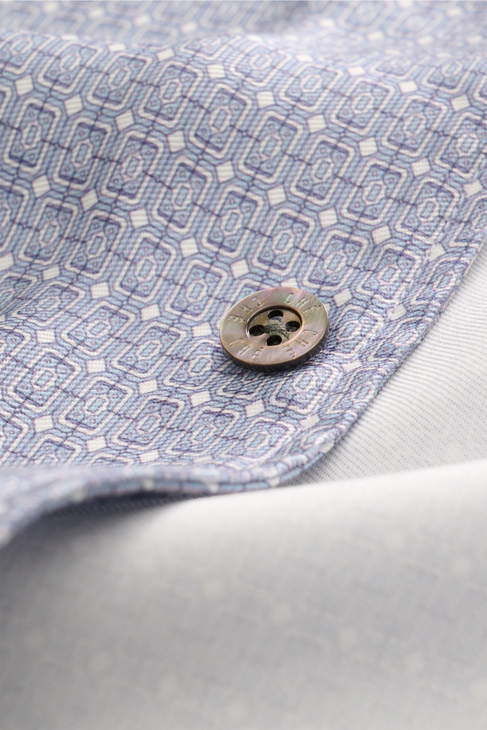 Close-up front view of the patterned fabric and button of the Ché Kurzarmhemd 'Lucena' Kubanischer Kragen rauchblau/offwhite gemustert short sleeve shirt. Das Kurzarmhemd 'Lucena' von CHÉ zieht mit ornamentalem Print, fließendem Lyocell und entspannter