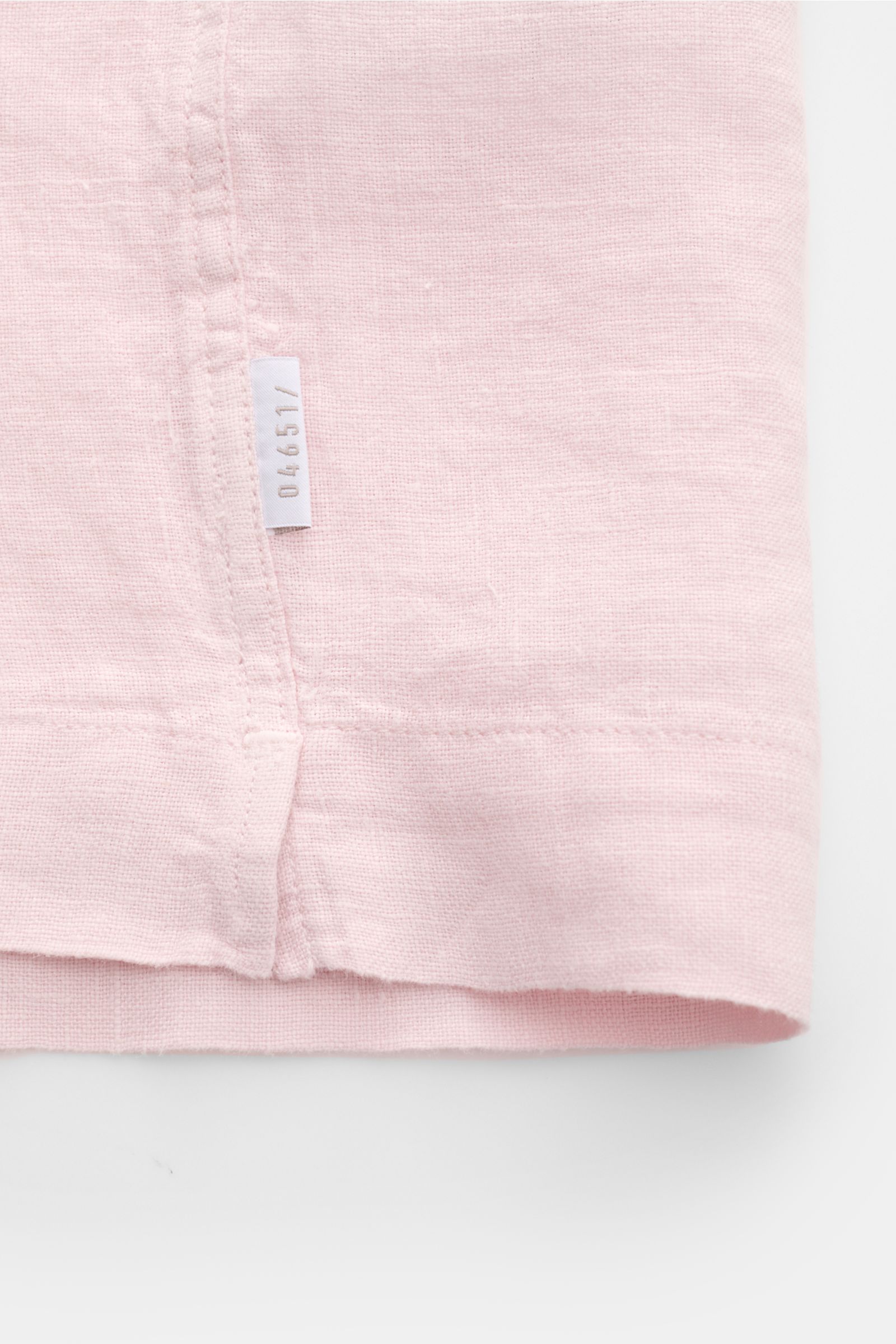 Nahaufnahme des Saumendes in roséfarbenem Leinen mit kleinem Weblabel 04651, Detail des 04651/ A trip in a bag Leinen-Kurzarmhemd Kubanischer Kragen rosé mit geradem Saum und kurzen Saumschlitzen, aus reiner Leinenqualität mit luftiger, materialtypischer 